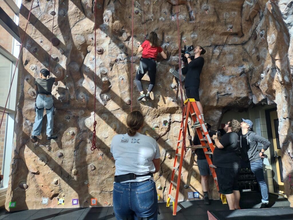 A film crew is recording a scene at an indoor climbing wall. Two people are climbing the wall, while a cameraman stands on a ladder filming one of the climbers. Several crew members are on the ground observing and assisting with the shoot. The setting is well-lit, and the climbing wall is equipped with ropes and climbing holds for different skill levels.