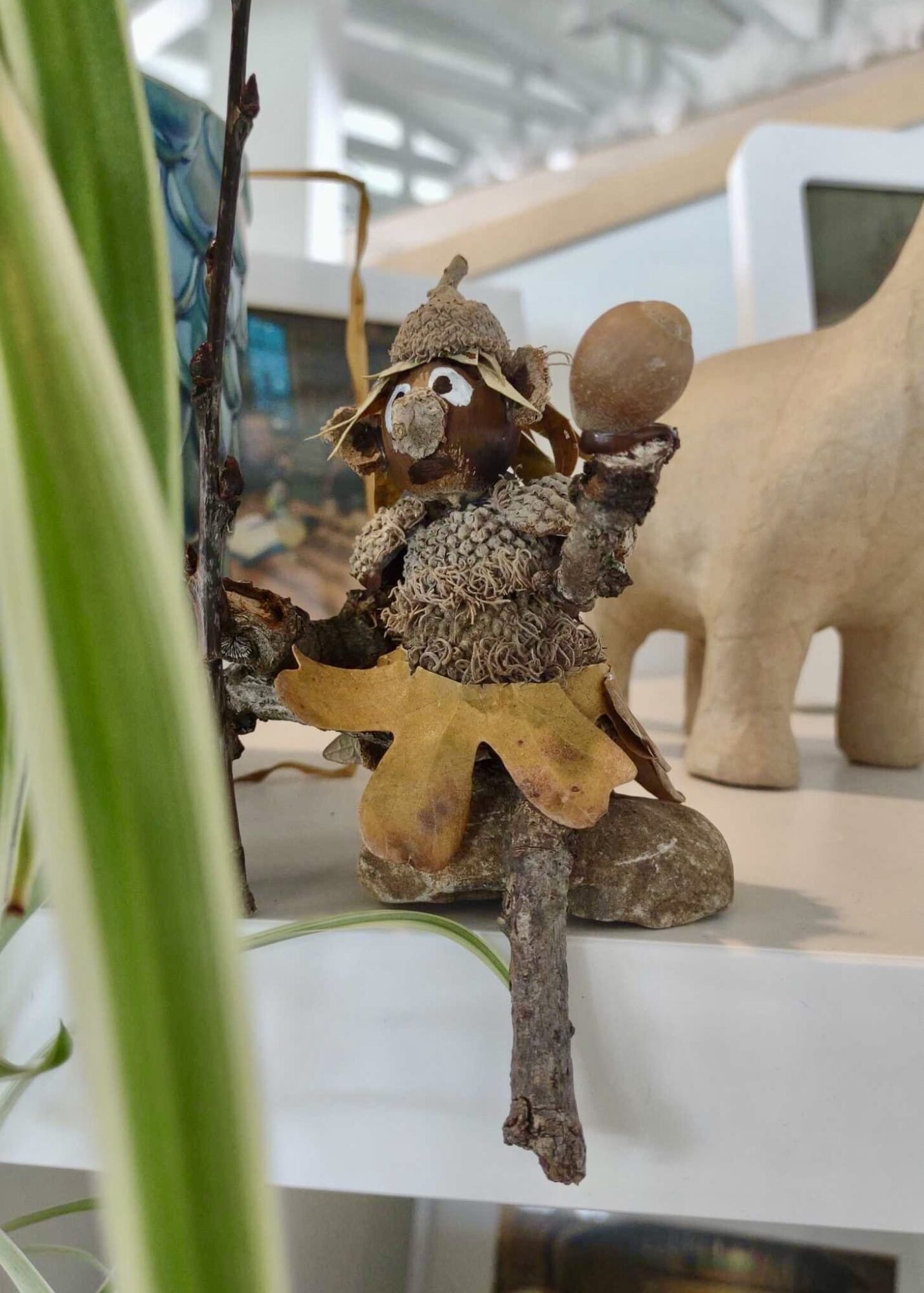 A small handcrafted figure made from natural materials such as acorns, twigs, moss, and leaves sits on a shelf. The figure has a whimsical appearance, wearing a leaf skirt and an acorn cap, with a twig arm raised holding an acorn. The background includes part of a plant and other decorative items.