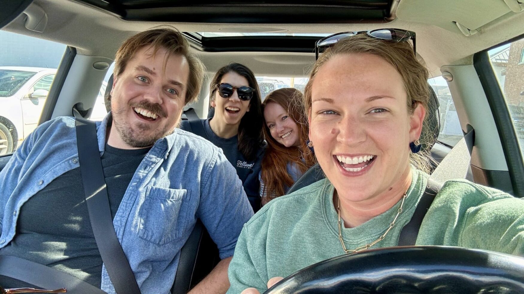Four people sitting in a car, all smiling and laughing. The two people in the front seats are a man in a denim shirt and a woman with light brown hair wearing a green sweater. In the back seat, there is a woman with long dark hair wearing sunglasses and a Tellwell shirt, and a woman with long red hair wearing a blue sweater.