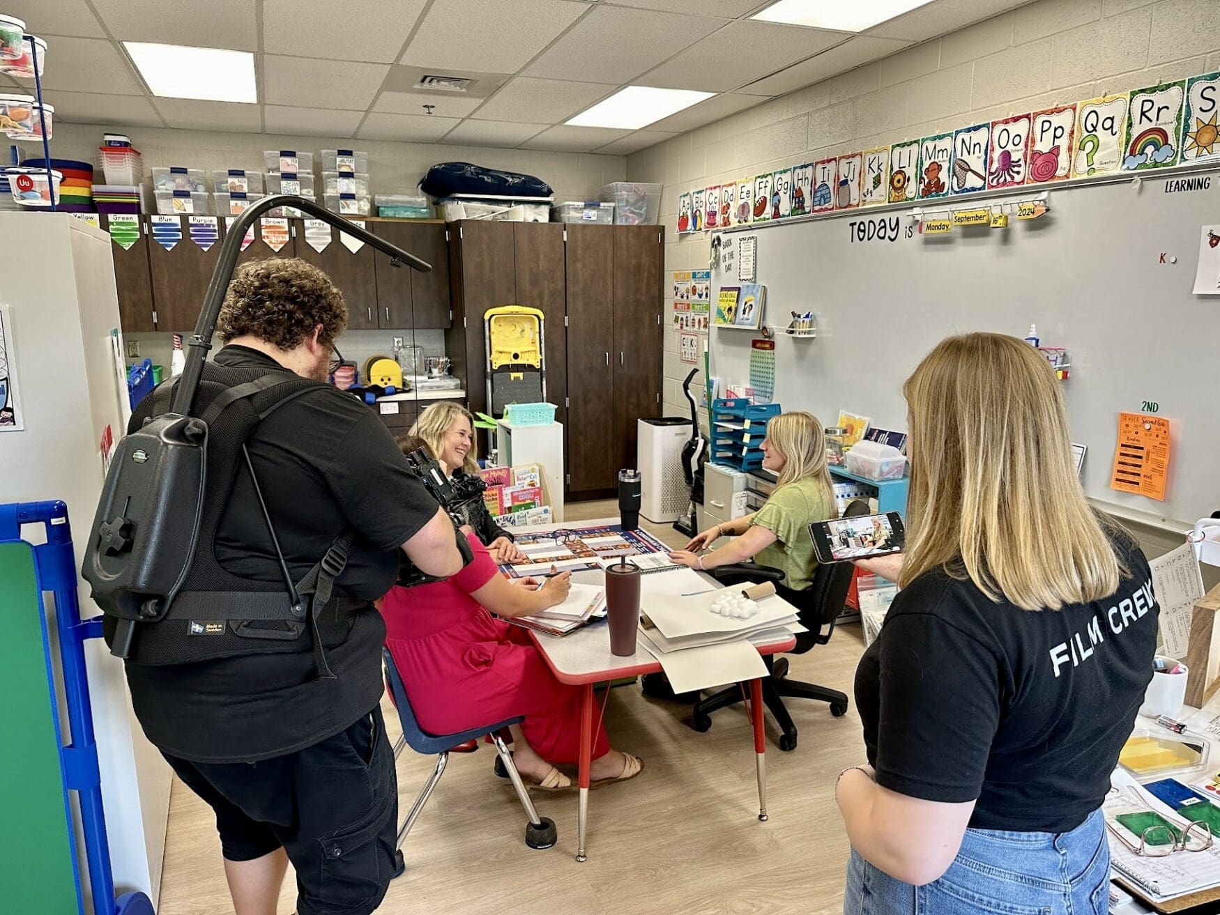 A cameraman wearing a body-mounted rig films two women seated at a table in a classroom while a 'Film Crew' member watches and monitors the footage on a phone. The women at the table are engaged in conversation and have papers and supplies spread out in front of them. The classroom is decorated with educational posters, including the alphabet and other children's artwork on the walls, and is filled with learning materials and storage bins.