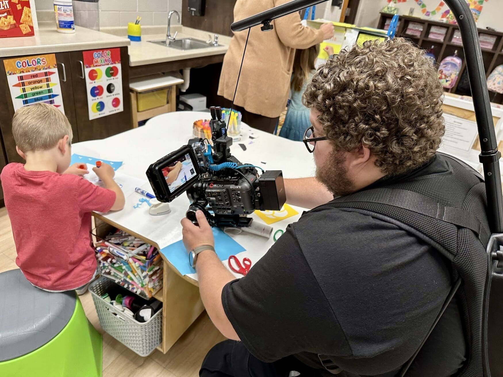 A cameraman wearing a body-mounted rig films a young boy working on an art project at a table. The boy, seated on a stool, is gluing paper as part of a craft activity. The table is filled with art supplies, including markers, scissors, and glue. The classroom walls display educational posters about colors, and various supplies and bins are visible around the room.