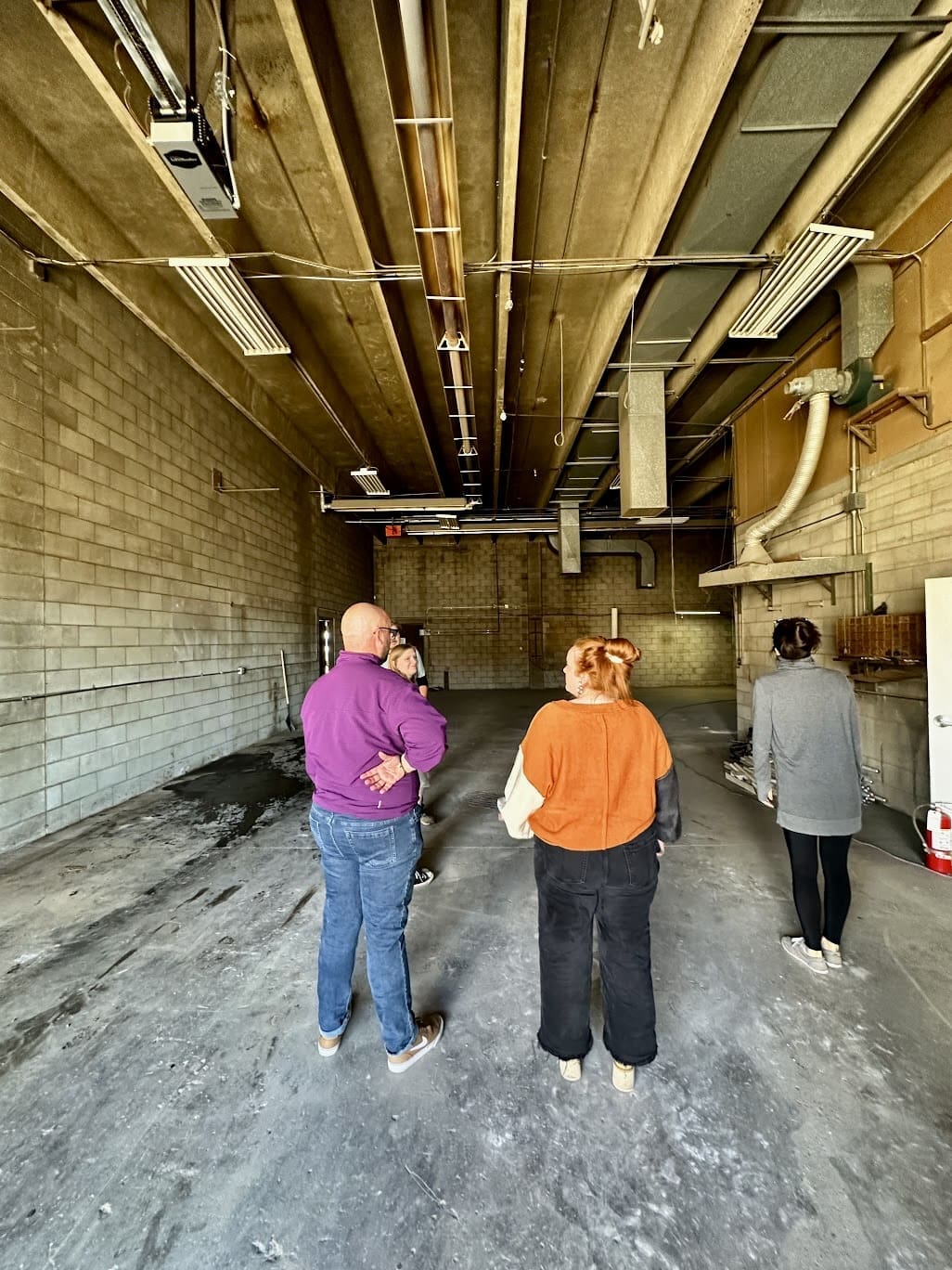 A group of four people stands in a large, industrial room with concrete walls and an exposed ceiling full of ducts and pipes. One person in a purple sweater appears to be leading the discussion.