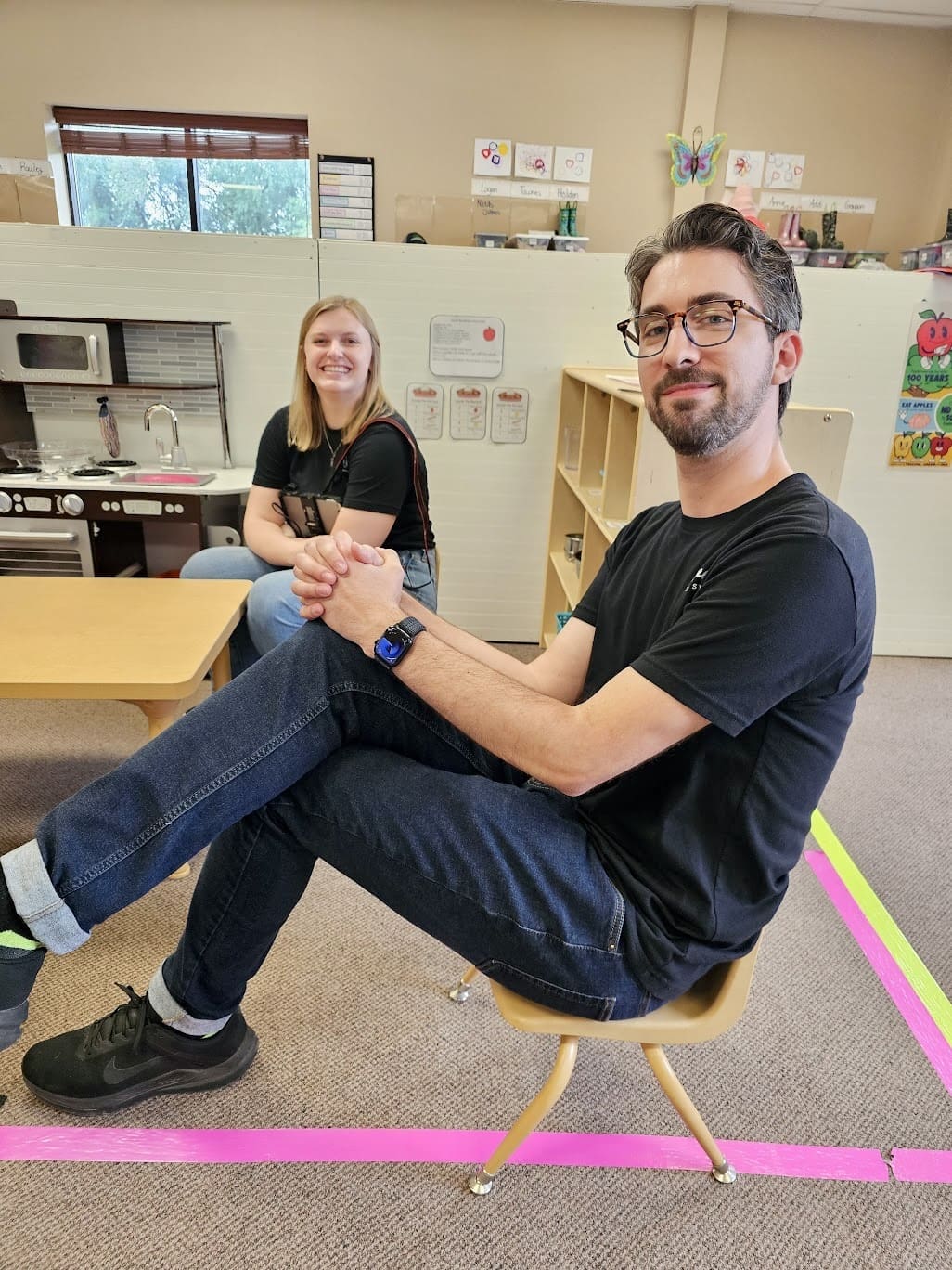 A man and a woman, both wearing black shirts, are seated in a brightly lit classroom. The man, sitting on a small chair with his legs crossed, smiles at the camera. The woman sits on a bench in the background, also smiling. The classroom features children's furniture, a play kitchen, and colorful decorations on the walls.
