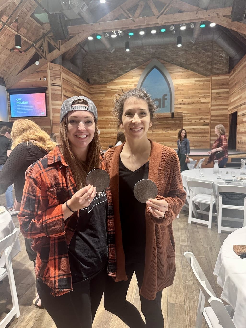 Two women standing inside a rustic venue with wood-paneled walls, smiling and holding round metallic coasters. One woman is wearing a backwards baseball cap, a black shirt, and a red flannel jacket, while the other is wearing a black shirt and a brown cardigan. In the background, there are people, tables set for an event, and a stage with a large arched window design.