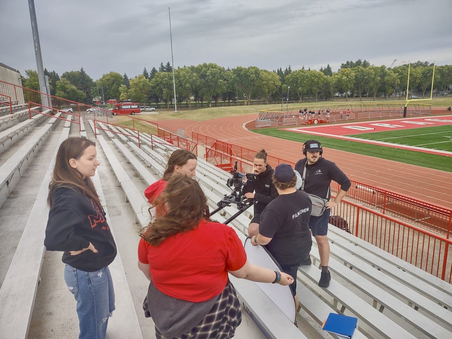 Group of film crew members setting up camera equipment on stadium bleachers, with two others observing. A red and white track and field is visible in the background, along with a 'Dragon Wagon van.
