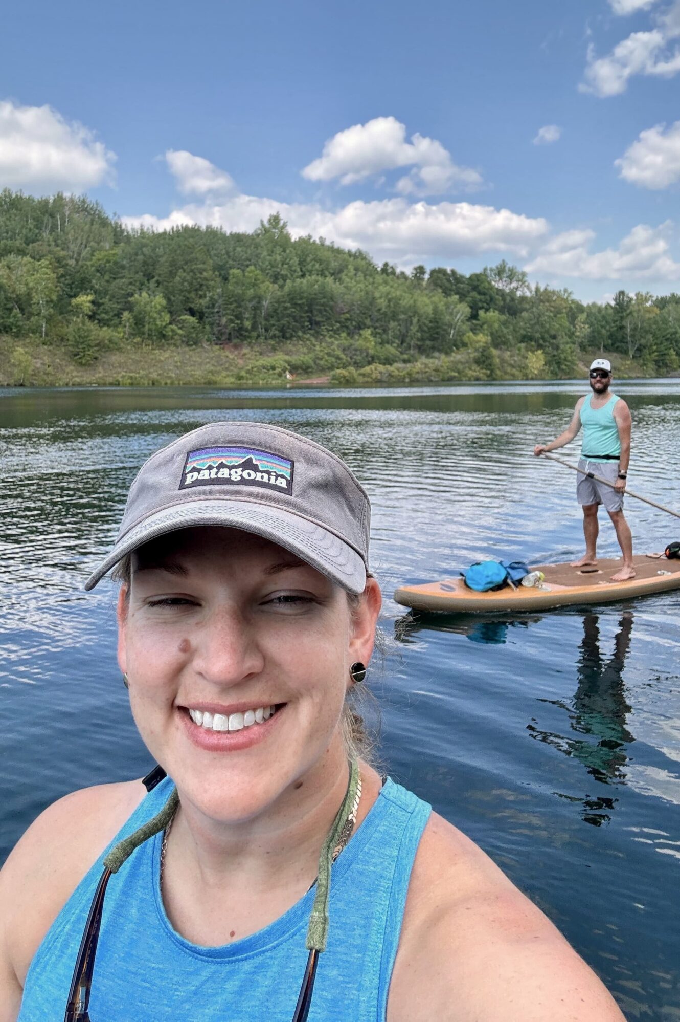 A woman in a blue tank top and gray Patagonia cap smiles at the camera while standing on a paddleboard on a calm lake. Behind her, a man in a blue tank top and sunglasses paddles on another board. The background features lush green trees under a bright blue sky with fluffy clouds.
