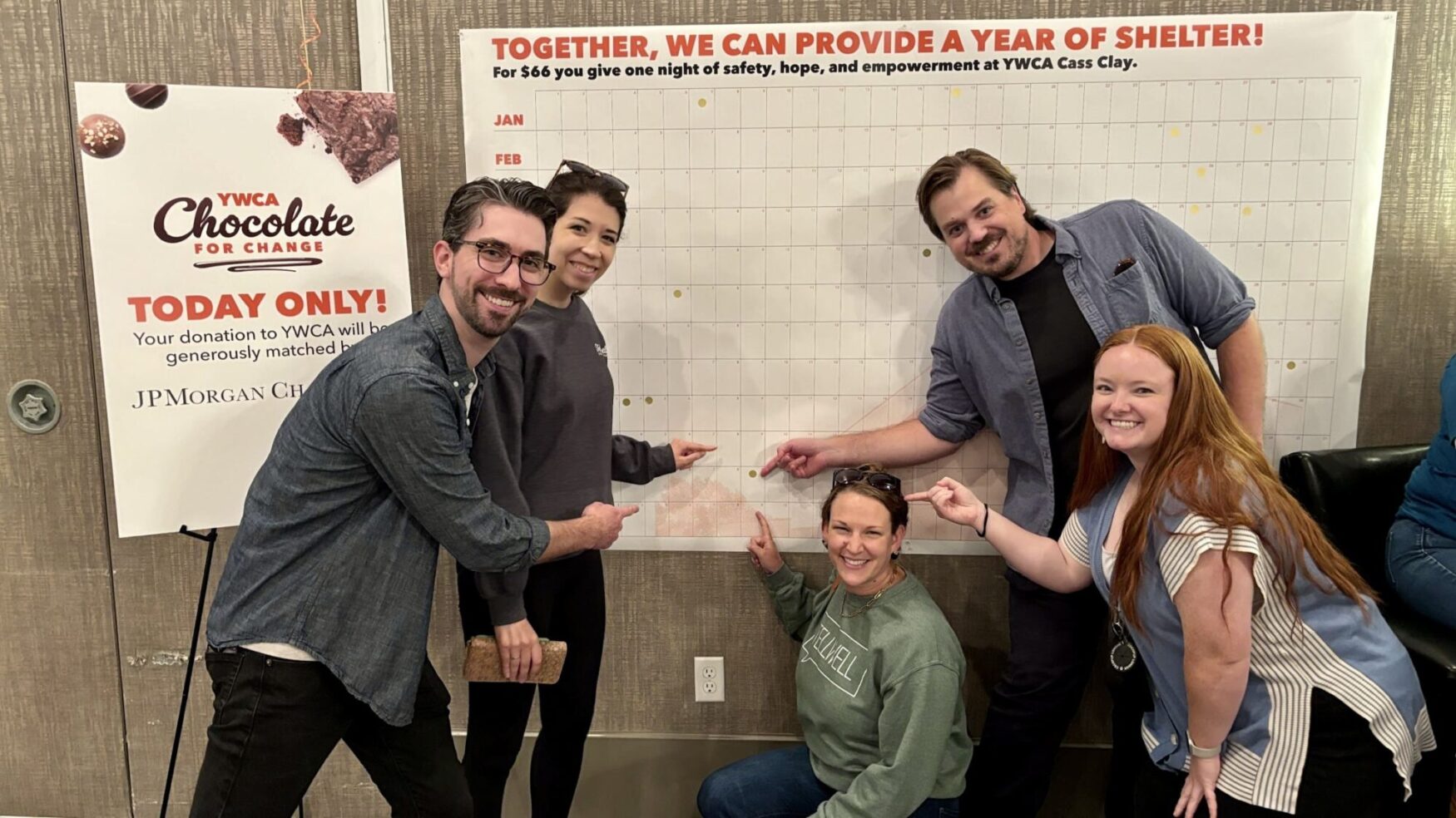 Five people smiling and pointing to different spots on a large donation tracking board for YWCA Cass Clay, which encourages donations to provide shelter. The board reads, 'Together, we can provide a year of shelter!' and mentions that $66 provides one night of safety, hope, and empowerment. There is also a sign on the left promoting YWCA's 'Chocolate for Change' event, with a 'Today Only!' message and sponsor logo from JP Morgan Chase.