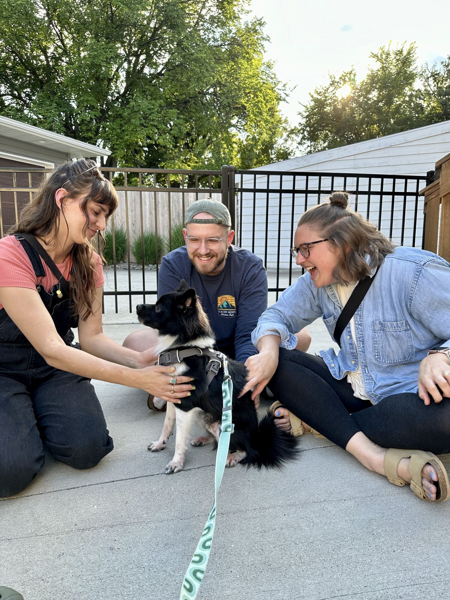 Three people sit on the ground outside, smiling and interacting with a small black and white dog wearing a harness and leash. The person on the left is gently petting the dog, while the others watch and smile. The scene is set in a backyard with trees and a fence in the background, under bright sunlight.