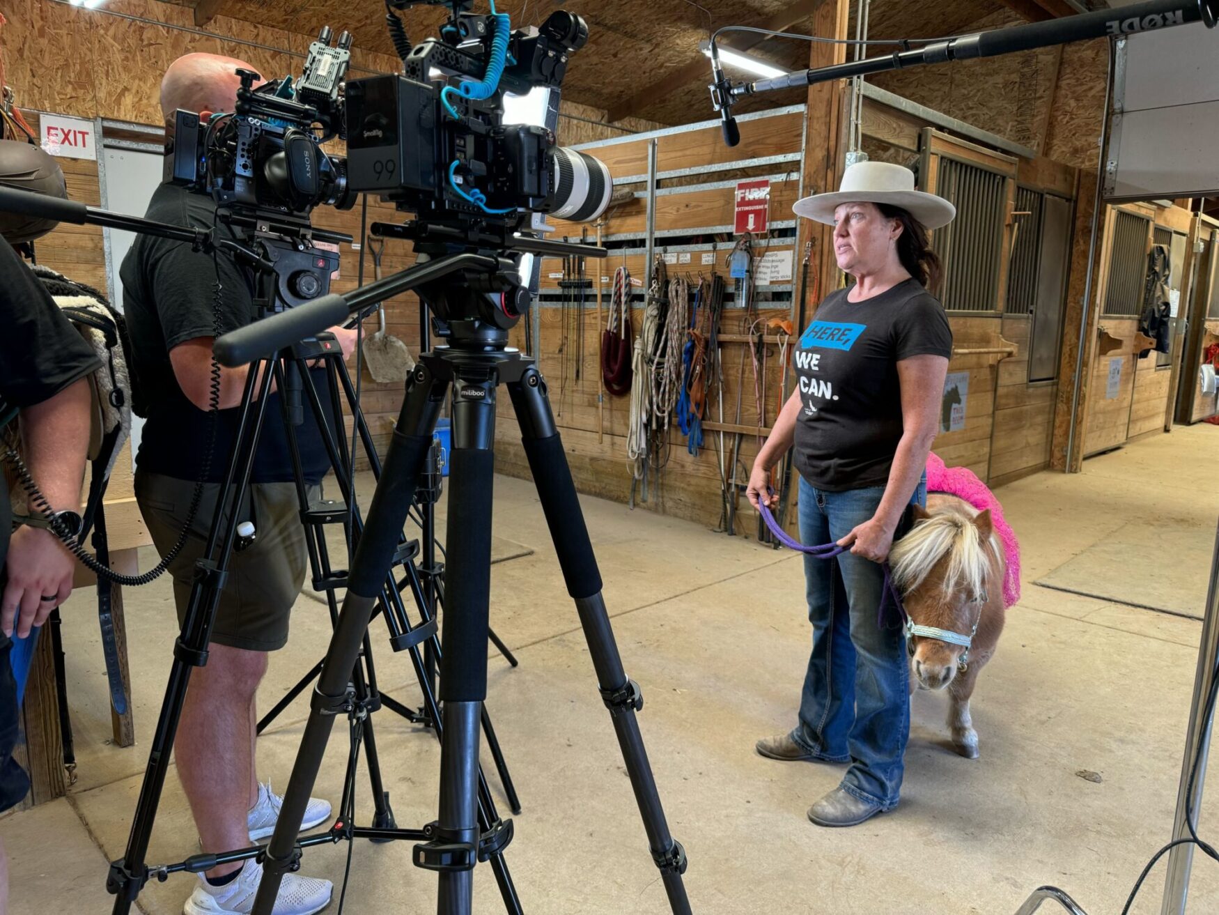 A woman in a wide-brimmed hat stands in a stable holding a small horse on a purple lead, with multiple cameras and a boom microphone set up around her for a film shoot.