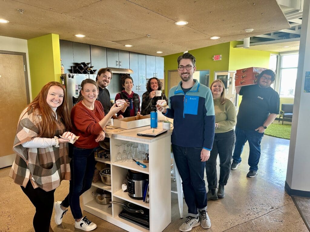 In this photo, a group of seven people is standing around a kitchen island in a bright, casual office environment, smiling and holding decorated cookies. A man at the back is carrying multiple boxes of donuts. The atmosphere looks cheerful and friendly.