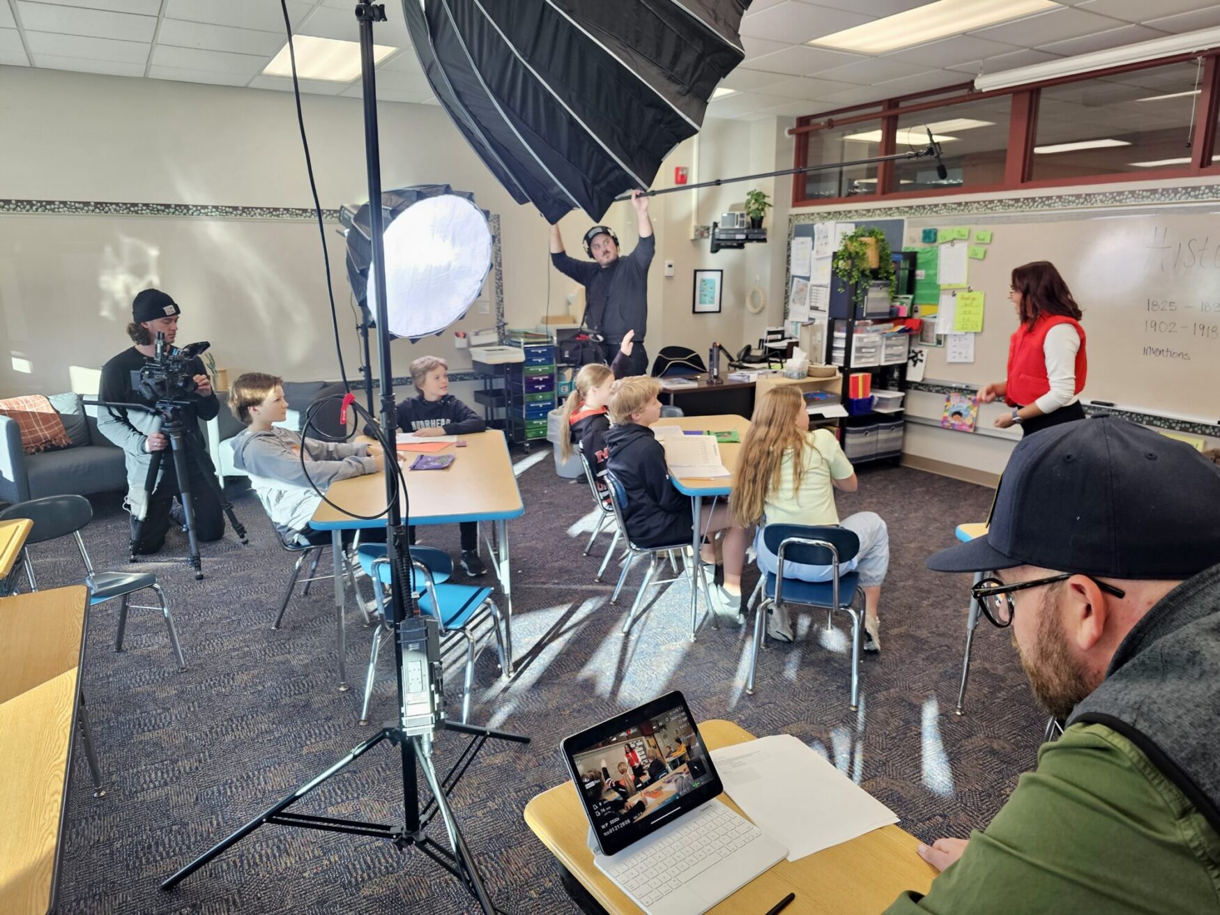 A classroom scene set up for a video shoot, with a group of children seated at desks facing a woman who appears to be their teacher. Several crew members are working around them: one person operates a camera, another holds up a large boom mic and light diffuser, and another watches the scene on a laptop in the foreground. The classroom is brightly lit, with various educational materials on the walls and desks.