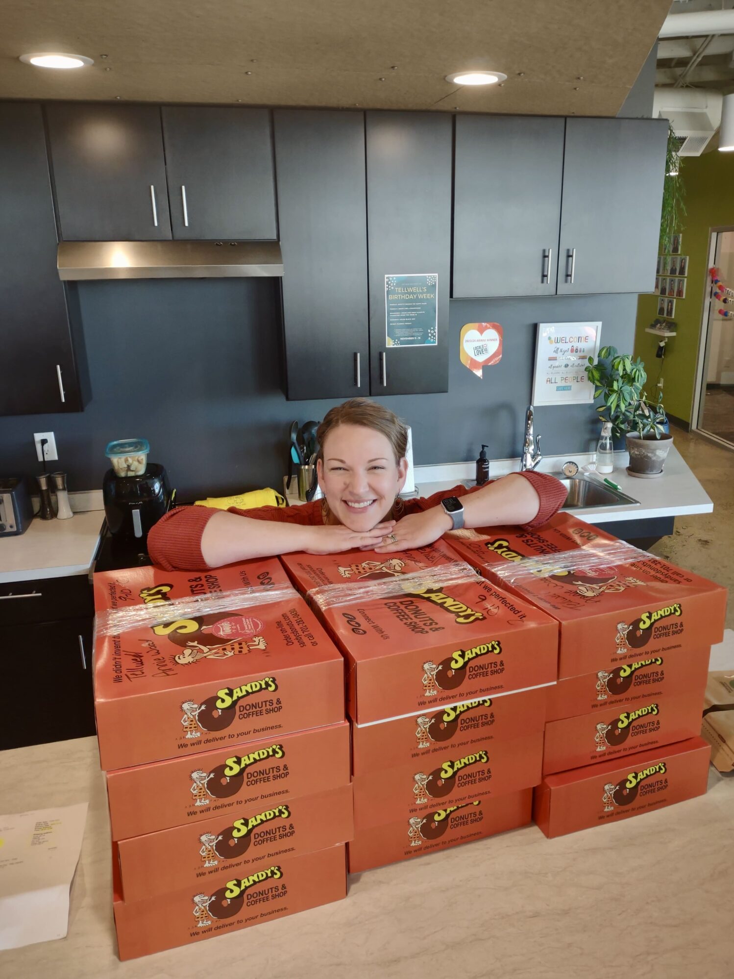 A woman is leaning her arms and chin on top of a stack of orange boxes from Sandy's Donuts in a kitchen area. She is smiling and posing behind about a dozen donut boxes stacked in two columns. The background shows black cabinets and some colorful posters on the wall, creating a fun and lively atmosphere.