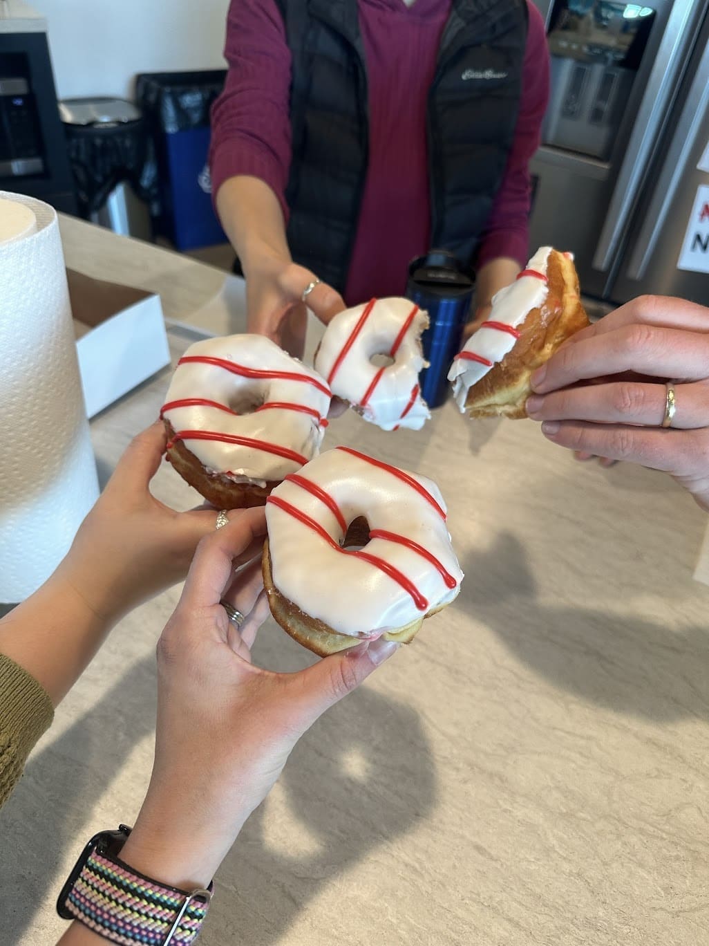 A close-up of four hands holding donuts with white icing and red drizzle, appearing to "cheers" them together in a celebratory gesture. The scene takes place on a light-colored countertop, and there’s a paper towel roll in the background, giving a casual, fun feel to the moment.