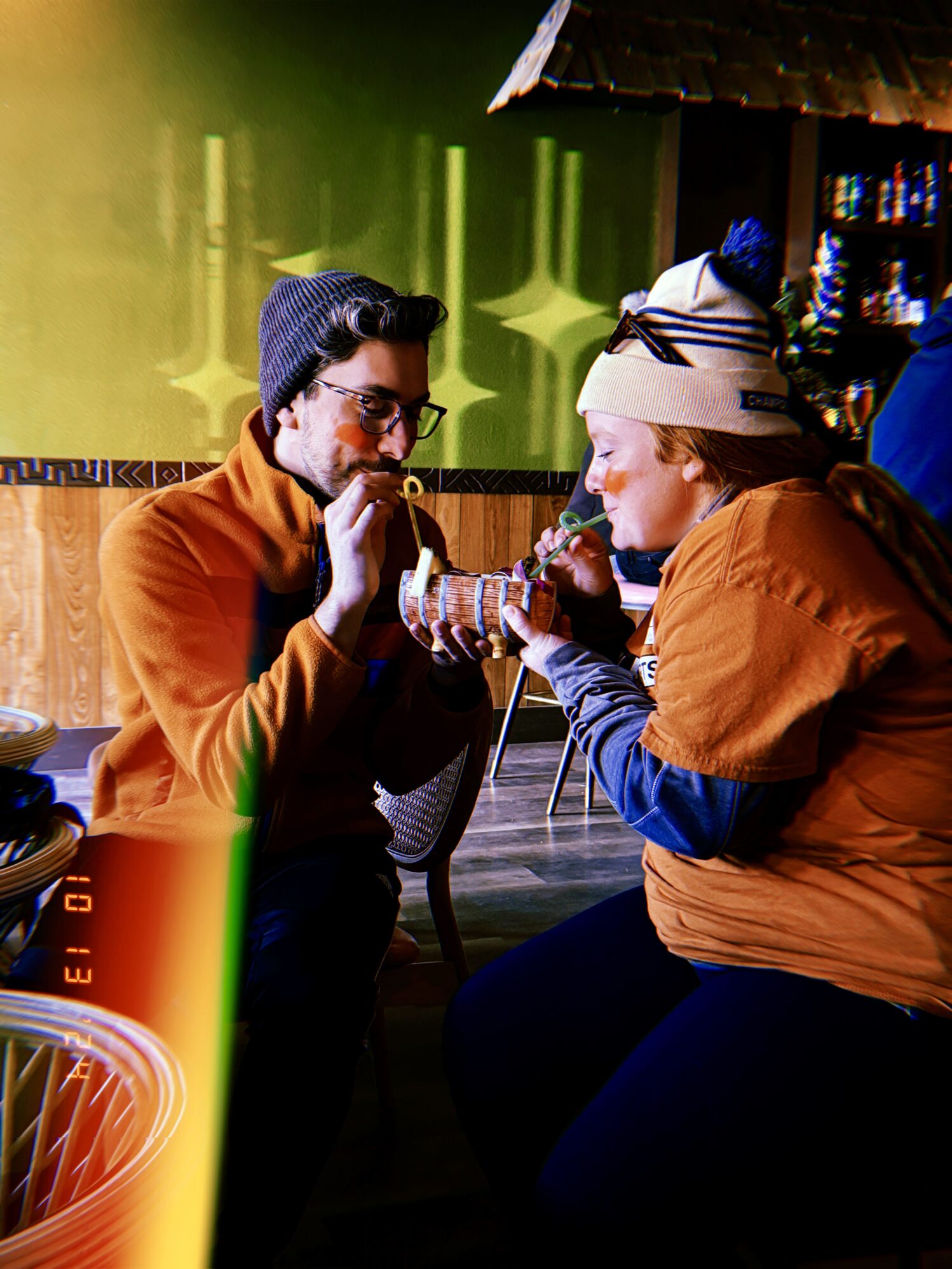A man wearing glasses and a winter hat shares a drink with a red haired woman wearing a winter hat. They both have orange face paint and matching shirts, signifying they are on a sports team. They are both using straws to drink out of a cup shaped like a wooden barrel. There is a filter on the photo, making it appear as if it is a film photograph. They appear to be in a trendy bar setting with tropical aspects to the decor.