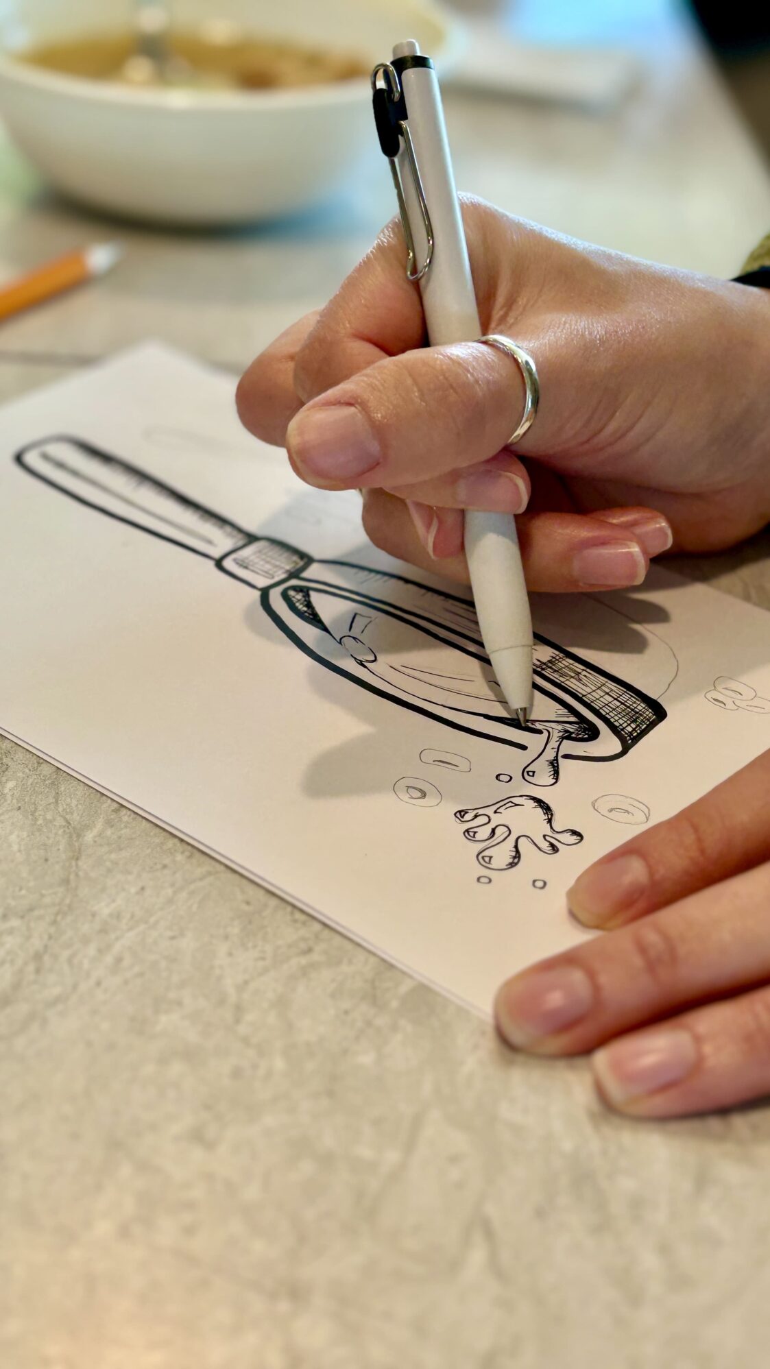 Close-up of a hand with a silver ring holding a pen, drawing a magnifying glass with liquid dripping from it. A bowl of food and a pencil are blurred in the background.