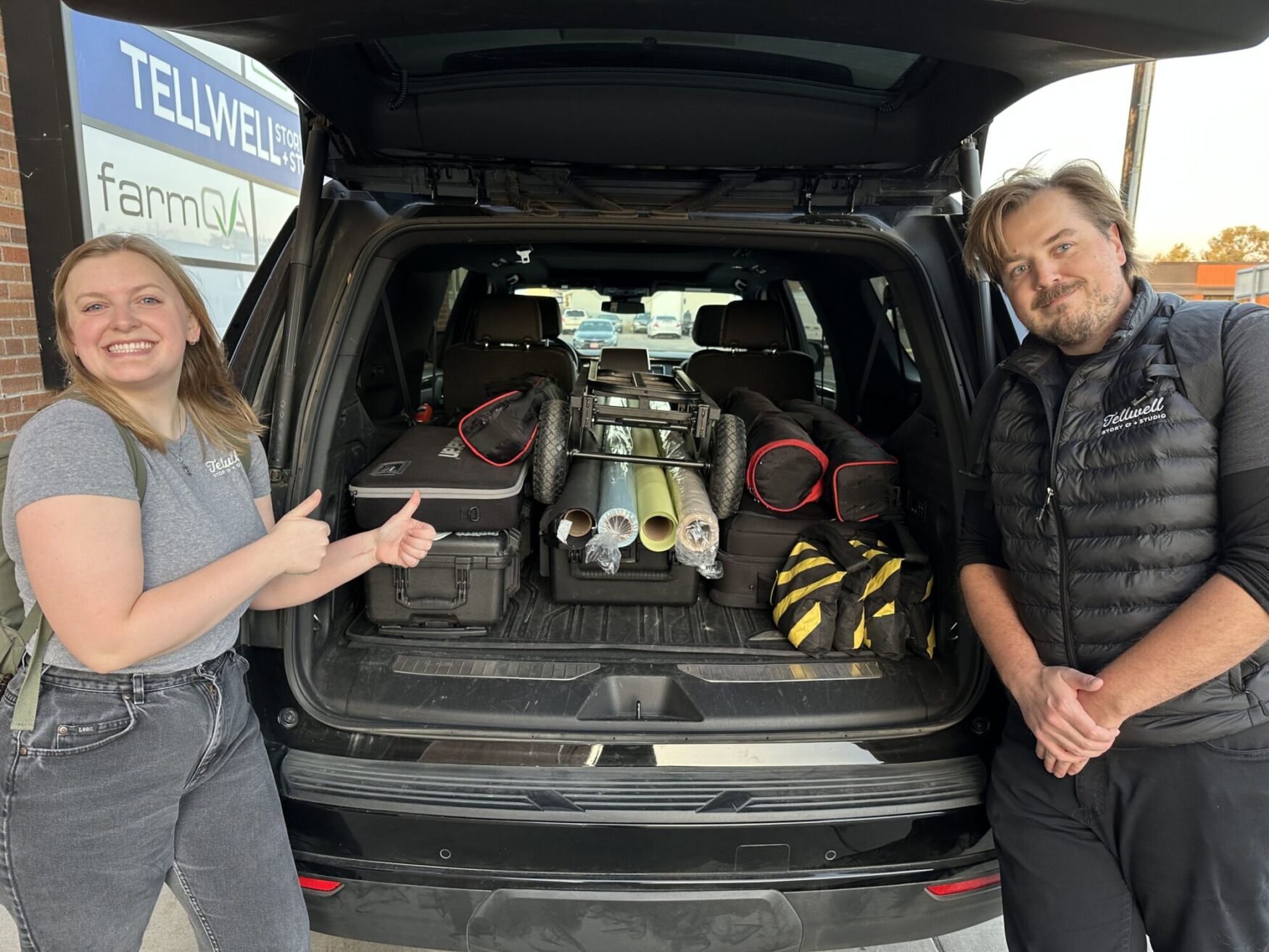A man and a woman in shirts that say “Tellwell Story Co. and Studio” standing in front of the trunk of a suburban. The suburban is filled with various film gear. The woman is smiling and giving two thumbs up and appears happy and proud. The man is holding his hands in front of himself and smirking with reassurance.