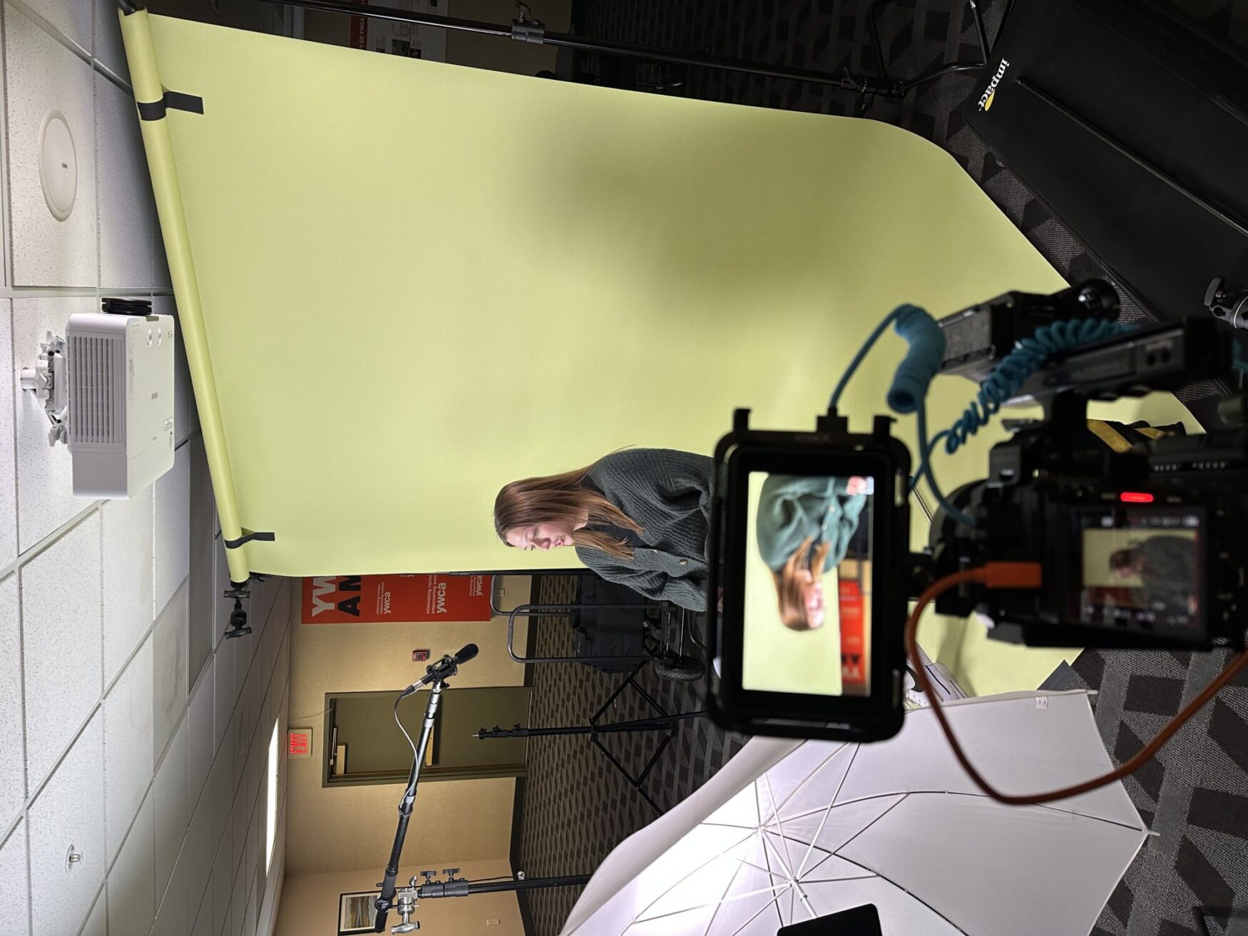 A woman is seated in front of a light green backdrop as part of a video production setup. She is facing a camera that is mounted on a tripod, with a microphone and a large umbrella light positioned nearby for optimal sound and lighting. The camera's display screen shows her image in focus as she prepares for filming. The setting appears to be an office or conference room, with "YWCA" signage visible in the background.