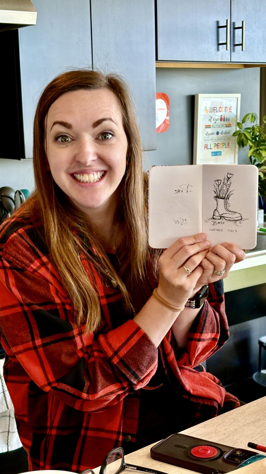 A woman in a red plaid shirt smiles while holding up a notebook with a drawing of a boot with flowers, labeled 'Boots' and 'Inktober Day 3.' She is seated at a kitchen counter with a phone and pens nearby.
