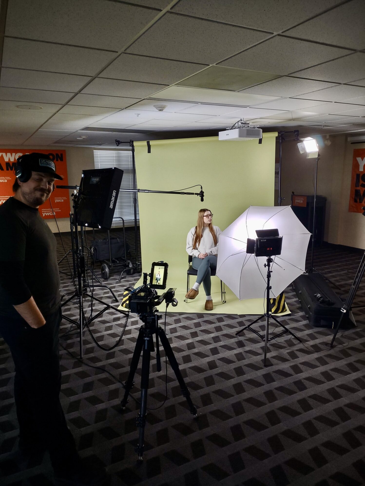 A woman is seated in front of a light green backdrop during a video shoot, with a camera and lighting equipment set up around her. A bright umbrella light and a softbox are positioned to illuminate her, and a microphone is hanging overhead on a boom arm. The camera on a tripod captures her image, while a crew member wearing headphones stands to the left, monitoring the scene. The background features "YWCA" signage, indicating the location is part of a YWCA facility.