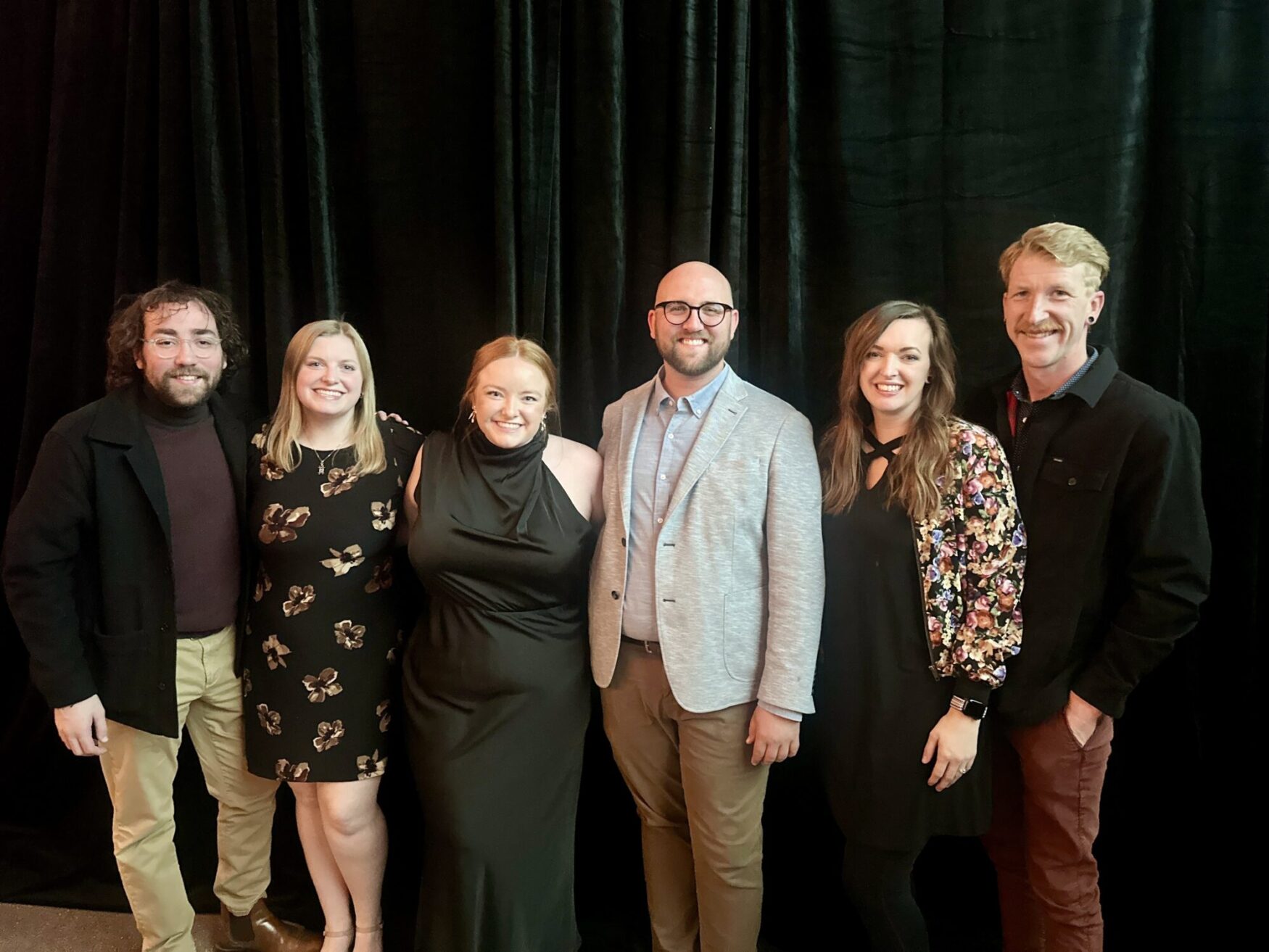 Six people stand in a line, smiling for a photo. They are all dressed formally, and standing in front of a black, velvet curtain. They appear to be at an event. There are three men and three women, intermixed within the line. They appear happy and friendly.