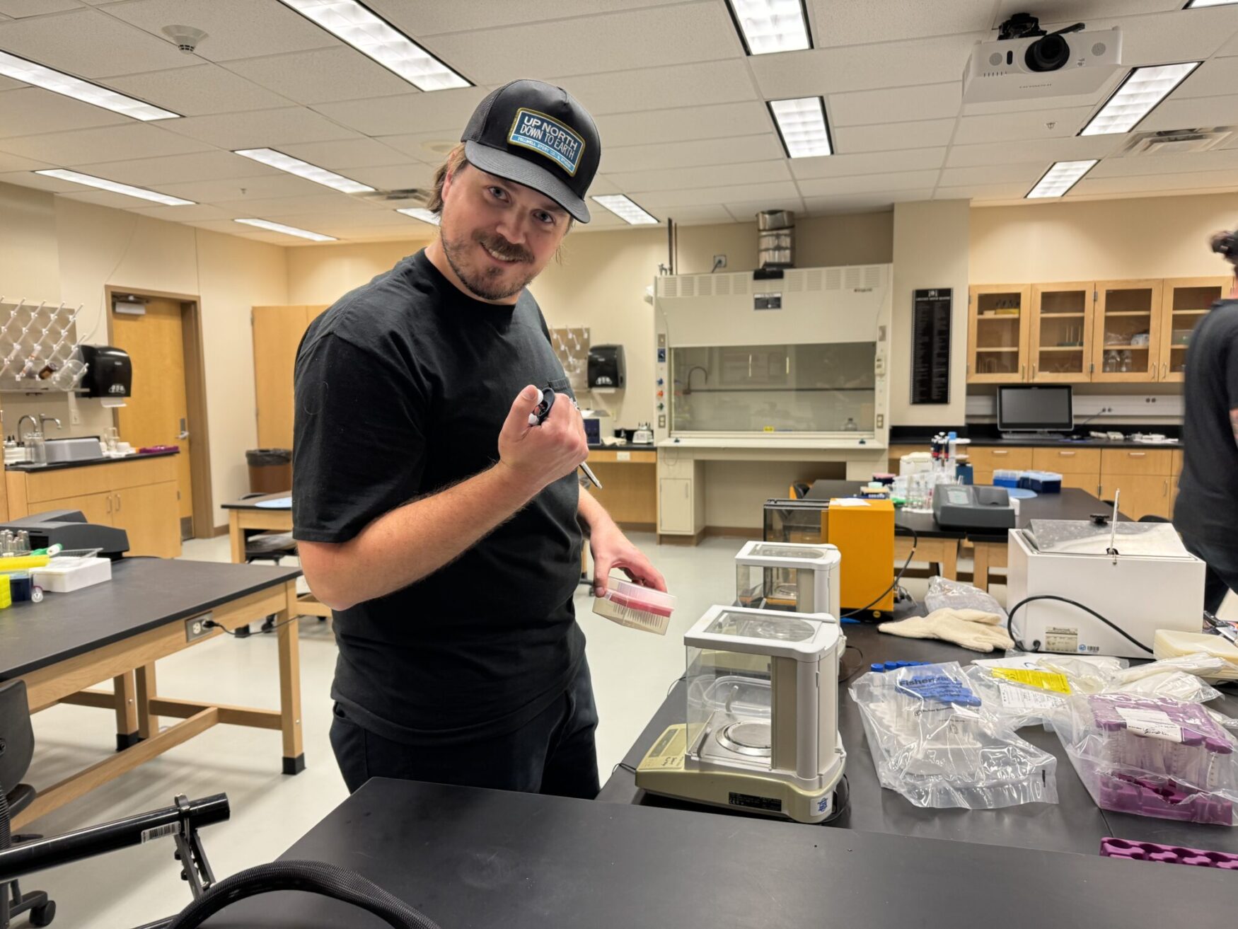 A man in a black t-shirt and a cap that reads "UP NORTH DOWN SOUTH" is in a laboratory setting. He stands at a table with a playful expression, holding up a lab tool, possibly a pipette. The table is filled with lab equipment, including scales, containers, and various scientific supplies. Behind him, there are shelves with glassware and other laboratory items, indicating an active science workspace.