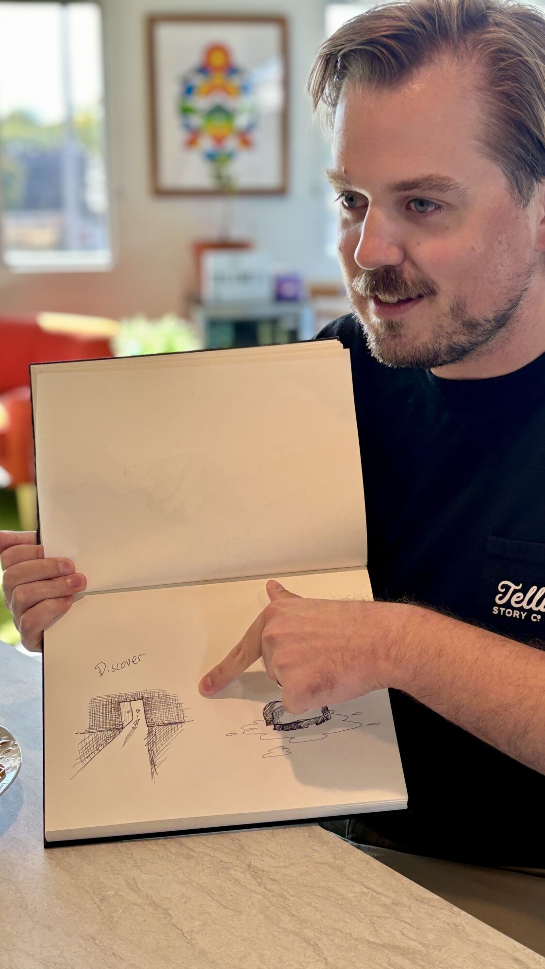 A man wearing a 'Tellwell Story Co.' t-shirt points at a drawing in an open sketchbook, featuring a tunnel leading to a door with the word 'Discover' and a sketch of an object floating on water. He is seated in a bright room with artwork in the background.