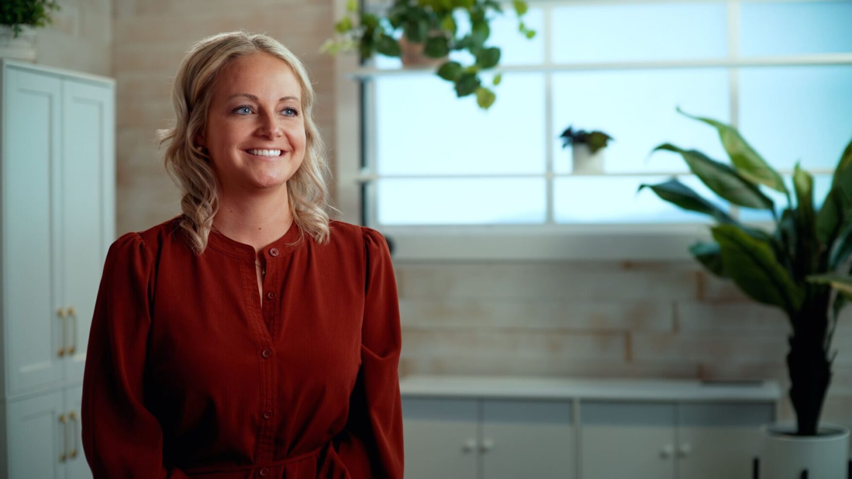 Smiling woman in a red blouse standing in a bright, modern office with large windows and green plants in the background.