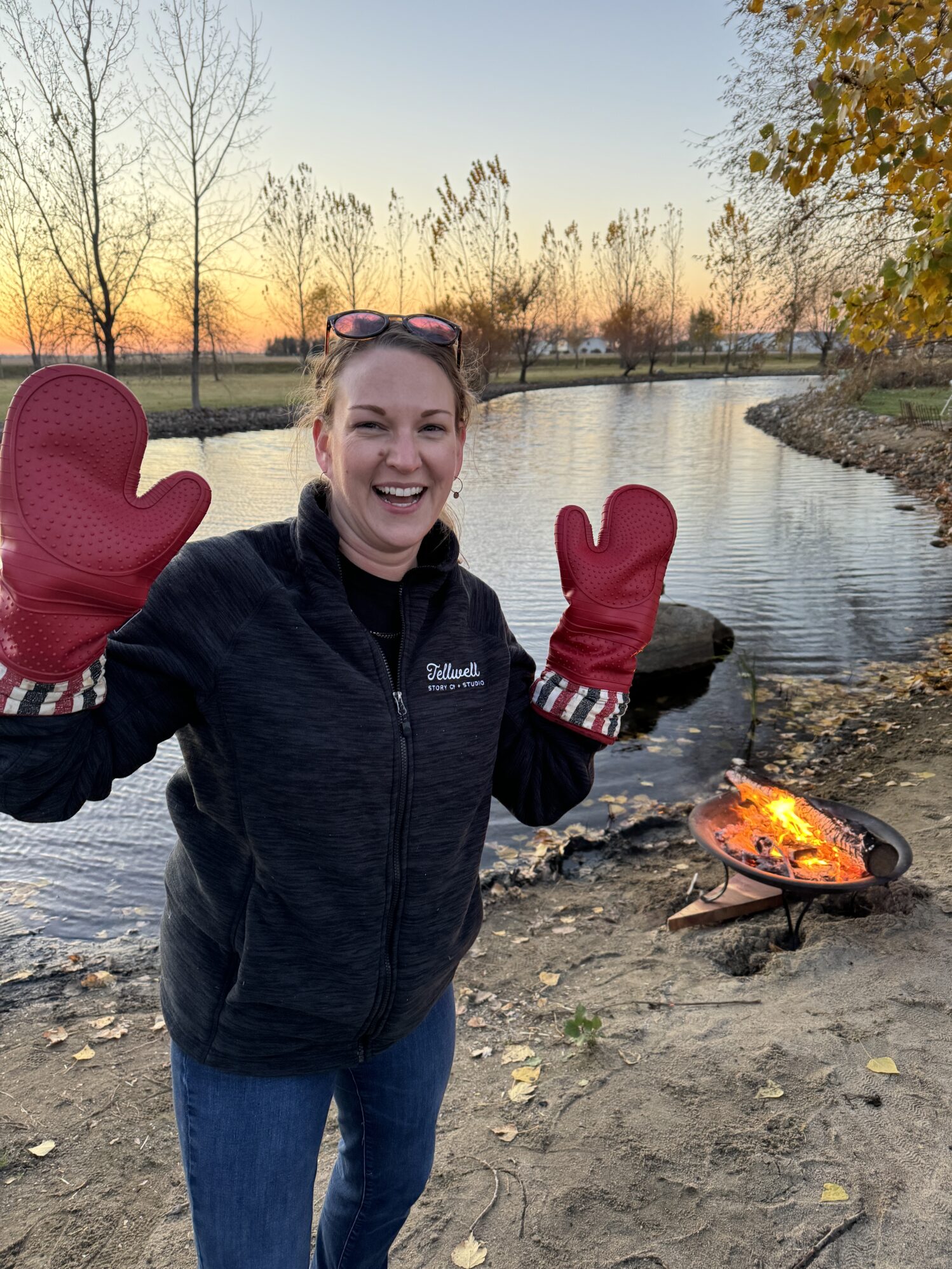 A smiling woman stands outdoors near a small fire pit by a peaceful river at sunset. She wears a dark jacket with "Tellwell" embroidered on it and large red oven mitts, playfully holding up her hands as if ready to handle something hot. The autumn leaves, calm water, and warm light of the sunset add a cozy atmosphere to the scene.