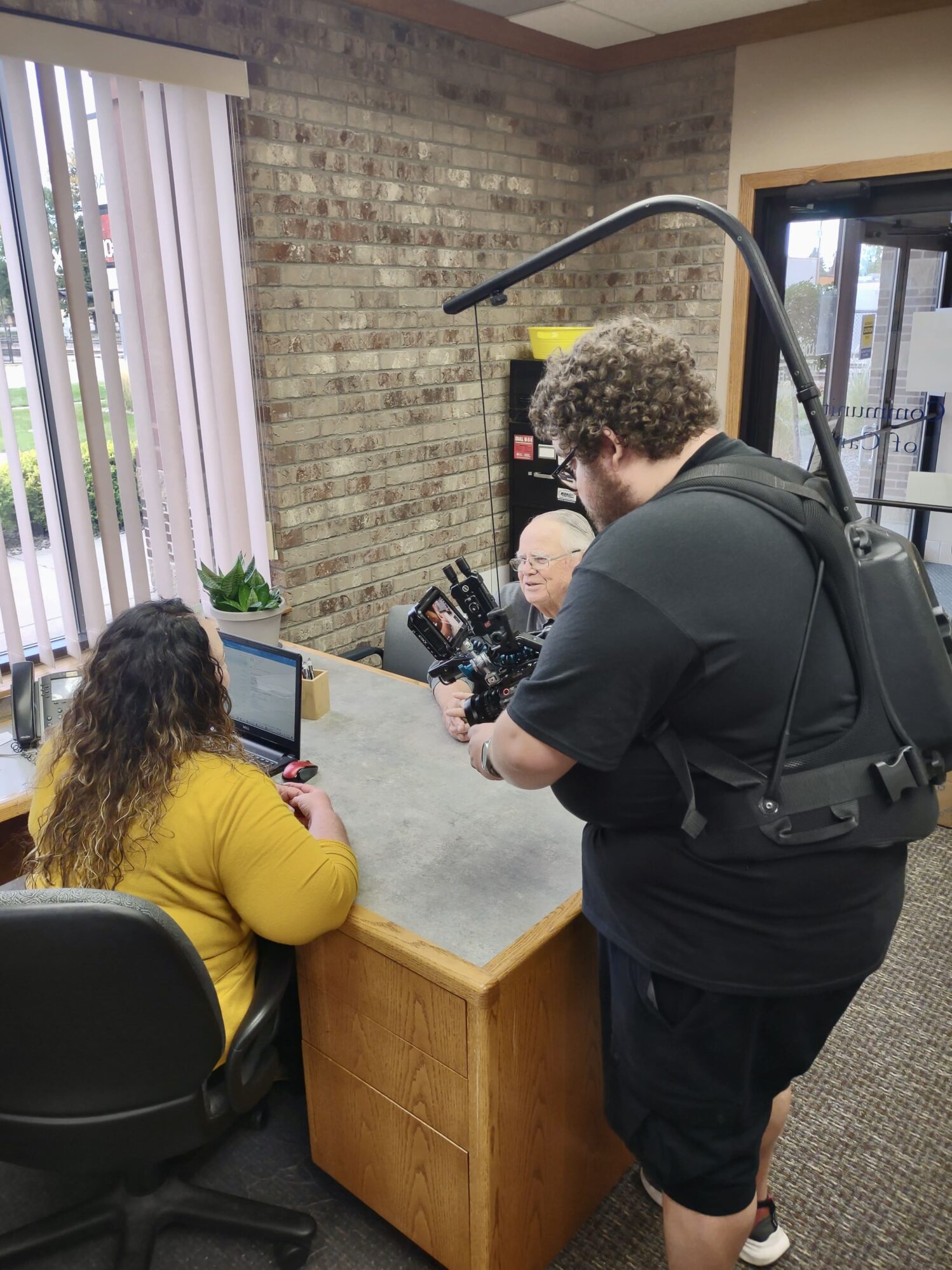A cameraman with a body rig films a woman in a yellow top working on a laptop at a desk, while an older man sits across from her in an office with brick walls.