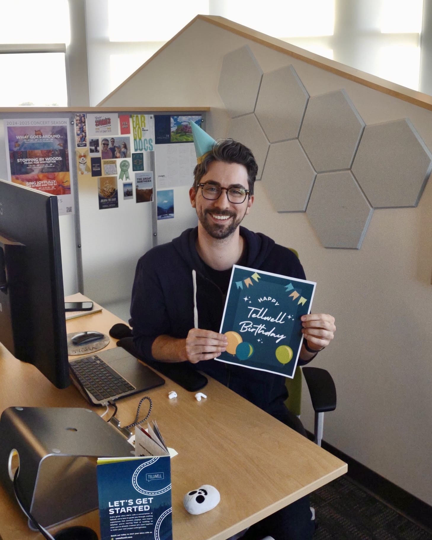A man with glasses and a beard sits at a desk in a cubicle of an office. There is a computer monitor on the desk along with a keyboard and a computer mouse. He is wearing a light blue birthday hat on the left side of his head and holding a paper that says “Happy Tellwell Birthday”. There is miscellaneous art and 7 gray hexagons on the walls behind him. He appears happy and cheerful.