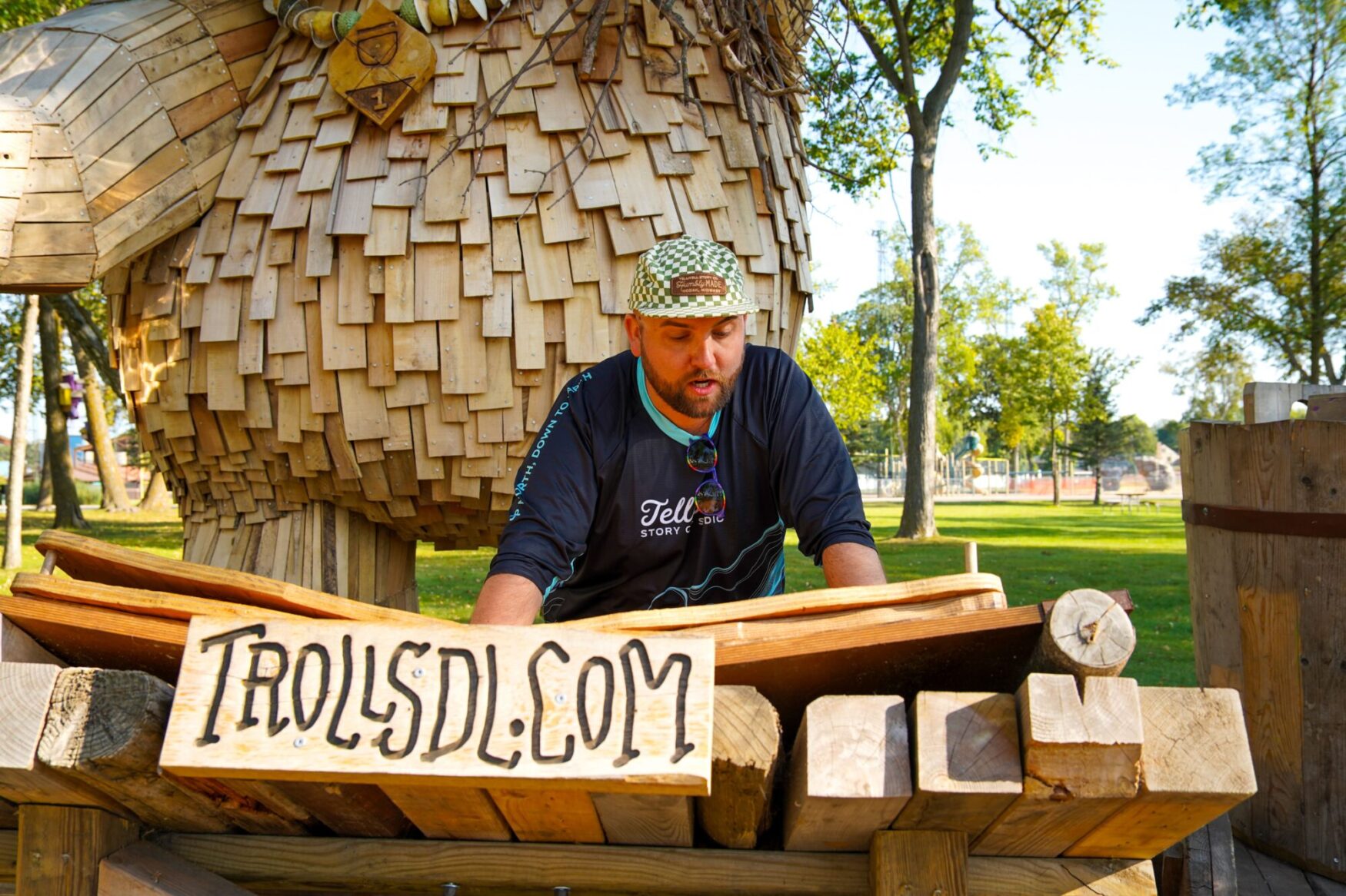 Person wearing a Tellwell Story Co. + Studio shirt and a checkered hat, standing behind a wooden sign that reads 'TrollsDL.com' in a park setting. The background includes a large wooden troll sculpture and trees.