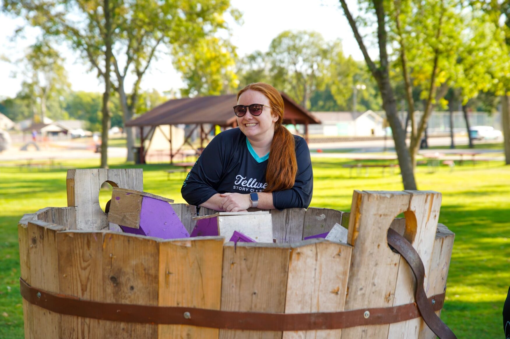 Person with long red hair and sunglasses, wearing a Tellwell Story Co. + Studio shirt, leaning on a large wooden structure with purple accents. The setting is a park with green grass, trees, and a pavilion in the background.