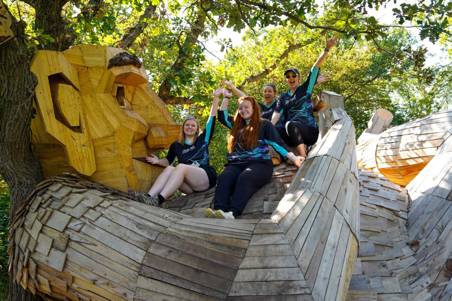 Group of people sitting on the arm of a large wooden troll sculpture, smiling and raising their hands in celebration. They are wearing Tellwell Story Co. + Studio shirts, surrounded by trees and enjoying a sunny day outdoors.