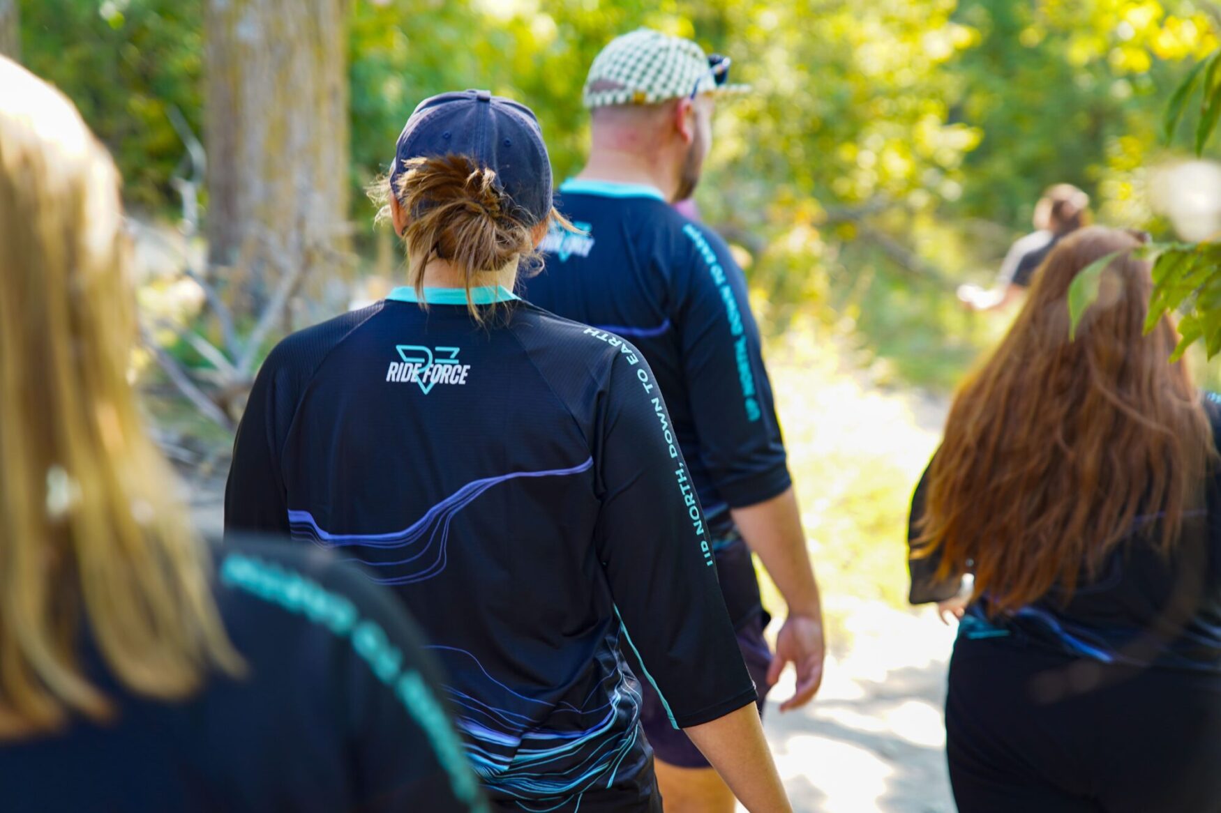 Group of people walking along a forest trail, seen from behind. They are wearing matching shirts with the 'Ride Force' logo, and the scene is surrounded by greenery in a sunlit outdoor setting.