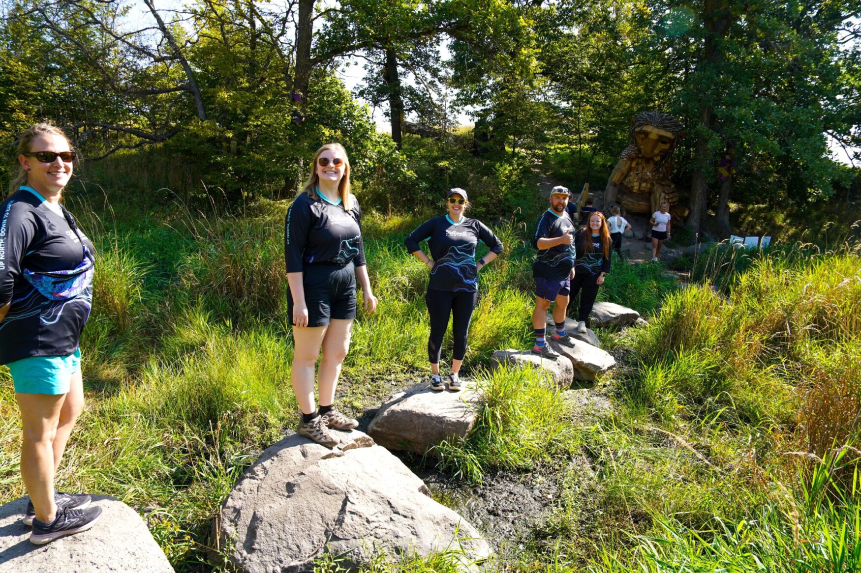 Group of people standing on large stones in a grassy area, with a wooden troll sculpture partially visible in the background. They are all wearing Tellwell Story Co. + Studio shirts and appear to be enjoying a sunny outdoor adventure.