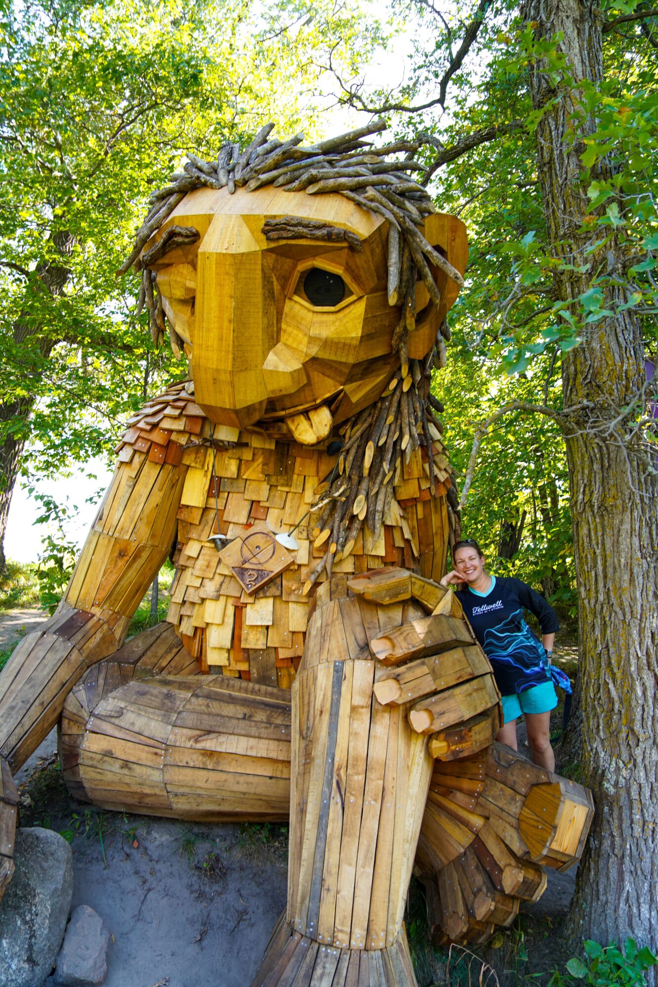 Large wooden troll sculpture sitting with one arm resting on its knee. A person wearing a Tellwell Story Co. + Studio shirt stands beside the troll, smiling. The scene is surrounded by green trees, creating a peaceful forest setting.