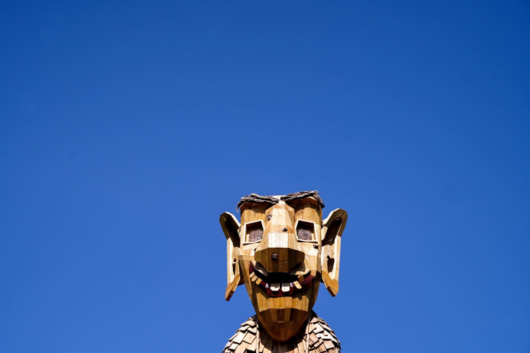 Close-up of a large wooden troll sculpture's head against a clear blue sky. The troll has a smiling expression, with large ears and square eyes, made from various pieces of wood.