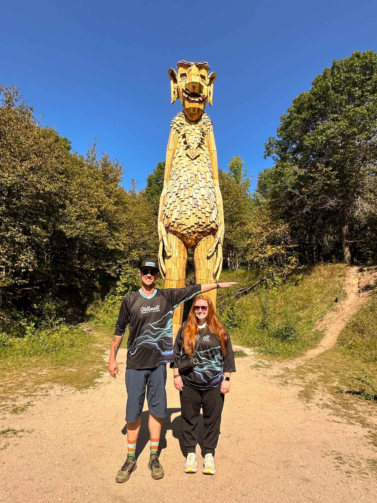 Two people wearing Tellwell Story Co. + Studio shirts stand in front of a tall wooden troll sculpture with arms extended, mimicking the troll's pose. The sculpture towers over them, surrounded by trees and set against a clear blue sky.