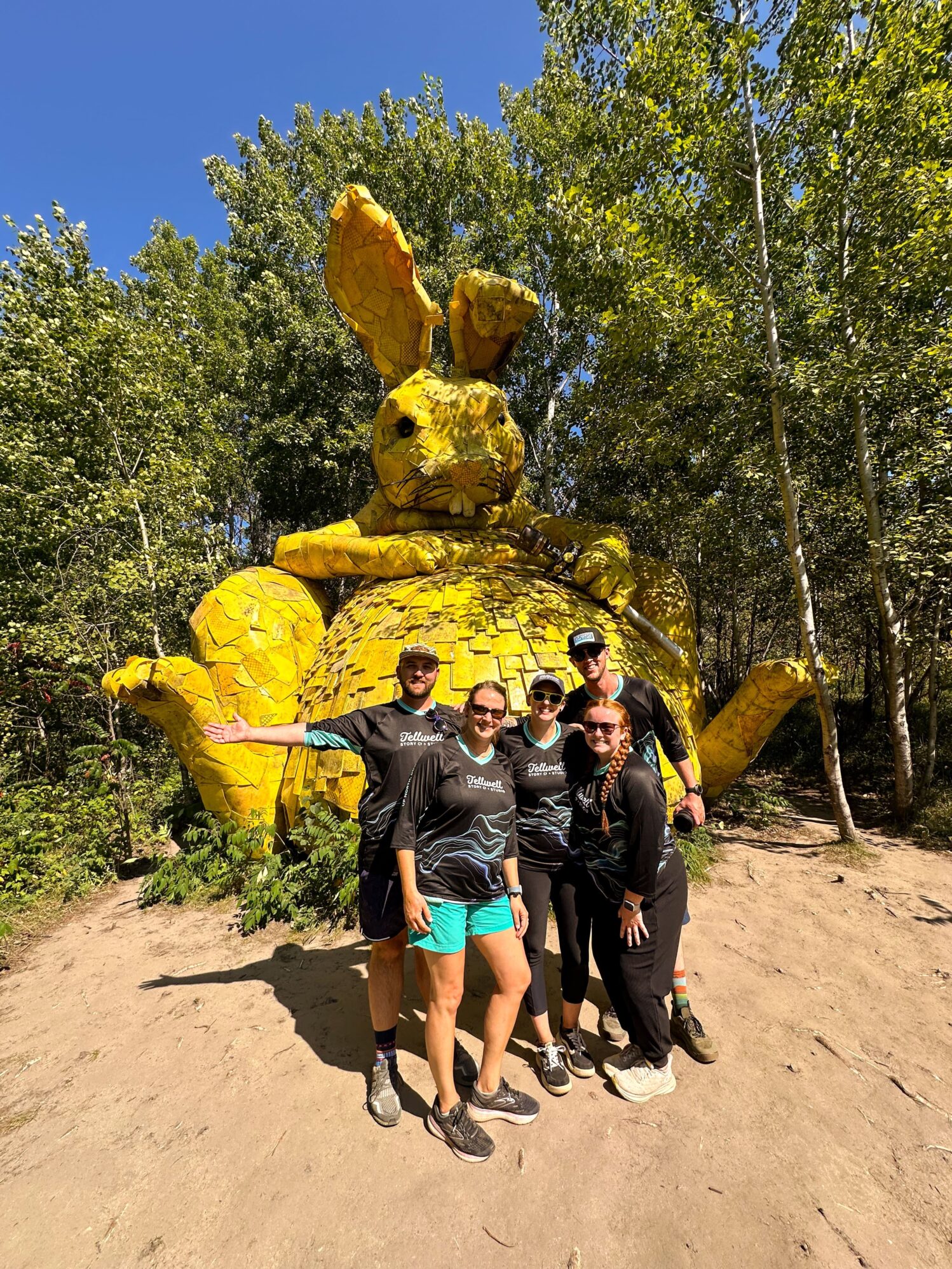 Group of people wearing Tellwell Story Co. + Studio shirts posing in front of a large yellow rabbit sculpture in a forest setting. The sculpture is made of textured material, and the group is enjoying a sunny day, surrounded by trees and greenery.
