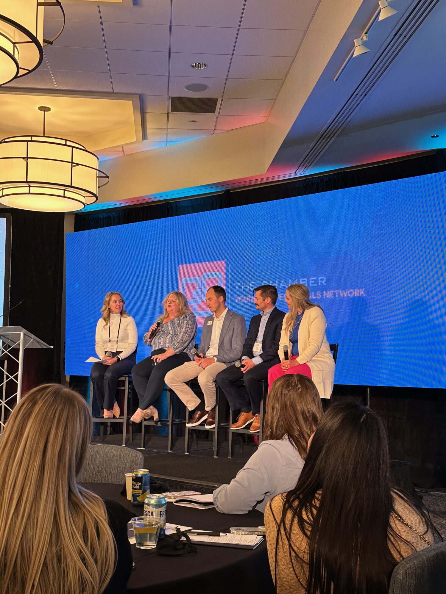 Panel of five speakers seated on stage at a conference, with one woman holding a microphone and speaking. The backdrop displays 'The Chamber Young Professionals Network.' Audience members sit at tables in the foreground.
