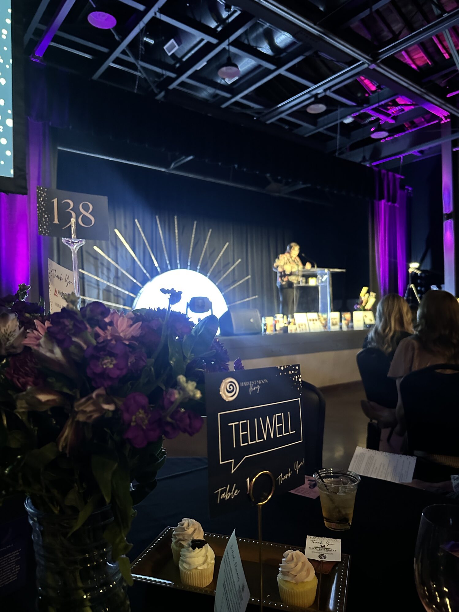 A vase of purple flowers, a plate of cupcakes, and a small sign that reads “Harvest Moon Fling Tellwell” with another smaller sign that reads “138” is shown on a table in the foreground. There is a woman standing on a stage in the background behind a pedestal. On stage, there is a large light fixture that is shaped like a sun. This appears to be at an event. The stage is filled with purple and pink lighting.