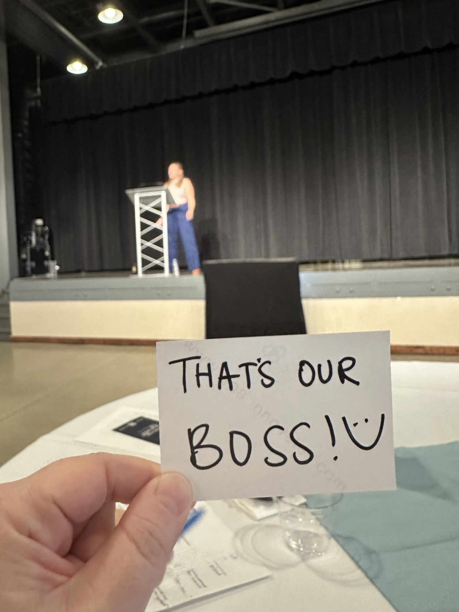 A hand holds up a small note card that reads “That’s our BOSS!” with a smiley face, as a woman speaks on stage in the background. The woman on stage is slightly out of focus, wearing a white top and blue pants, standing in front of a black curtain.