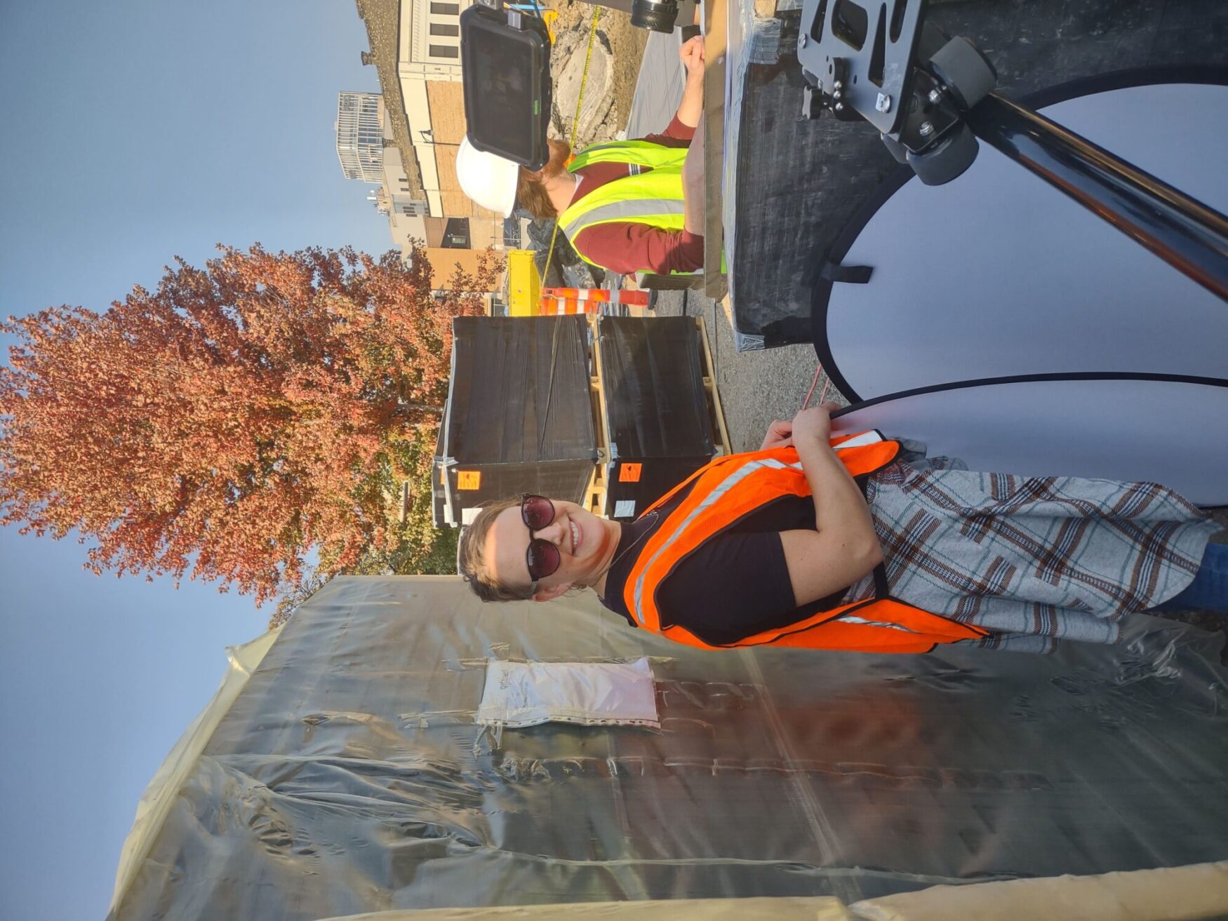 A woman in an orange construction and t-shirt vest handling equipment of film screens stands outdoors smiling at the camera, with a camera on a tripod in the foreground. A tree with orange leaves and a bright blue sky in the background.