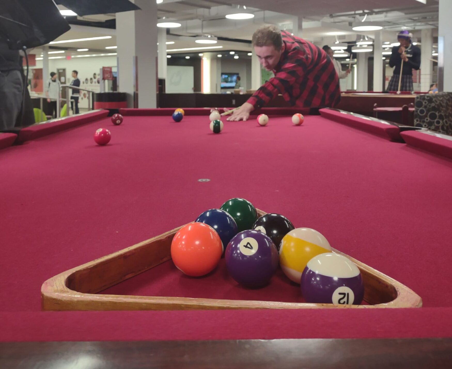 A young man at a billiards table preparing to take a shot of the cue ball in the background, with a handful of billiard balls in the wooden triangle in the foreground, all while on a red felt table.
