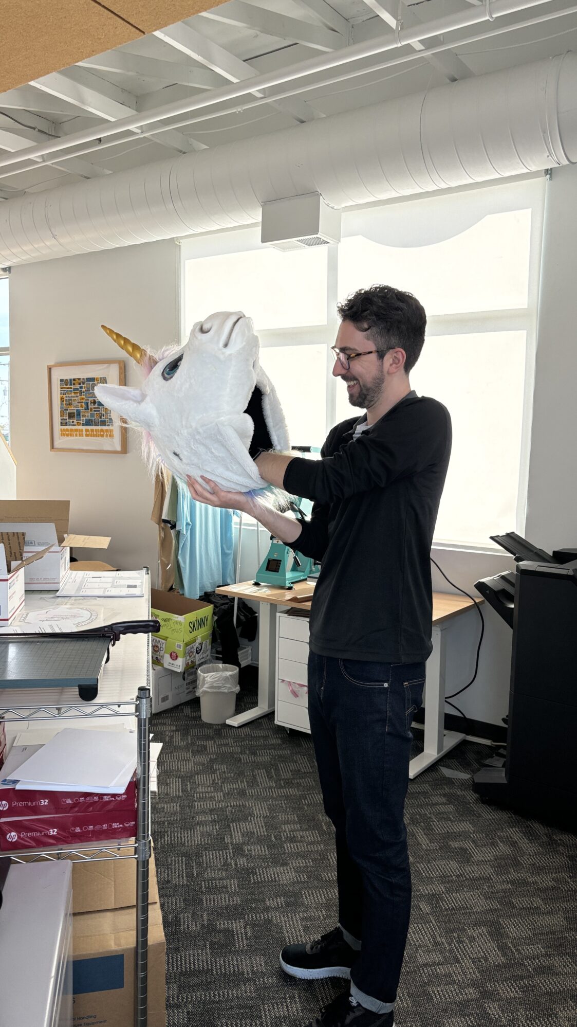 A man wearing glasses and a black long-sleeve shirt holds up a large white unicorn head prop, smiling as he examines it. The office space around him is bright with large windows and various office supplies in the background.
