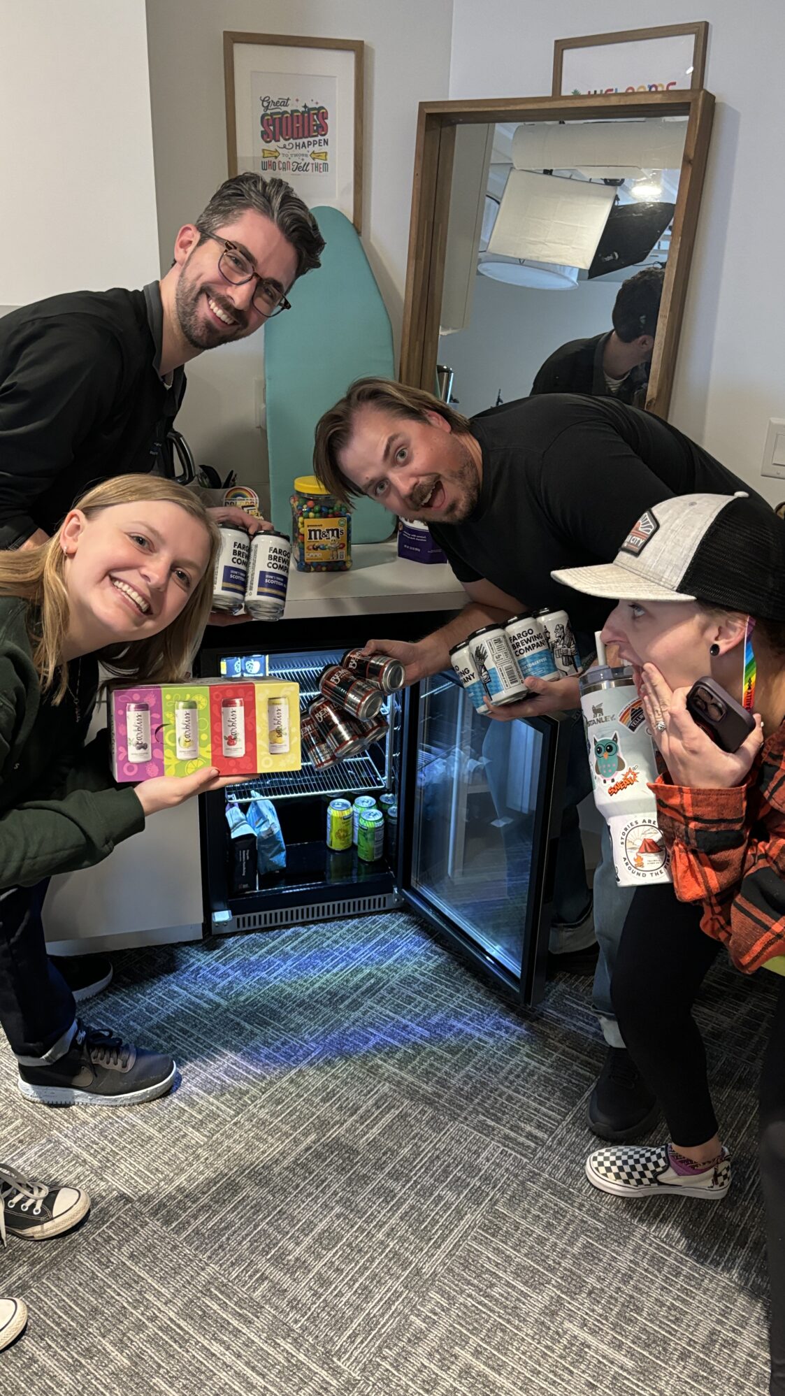 Four coworkers excitedly pose in front of a small fridge filled with drinks. They are holding cans of sparkling water and beer, smiling toward the camera. Behind them is a colorful office space with snacks and decorations.
