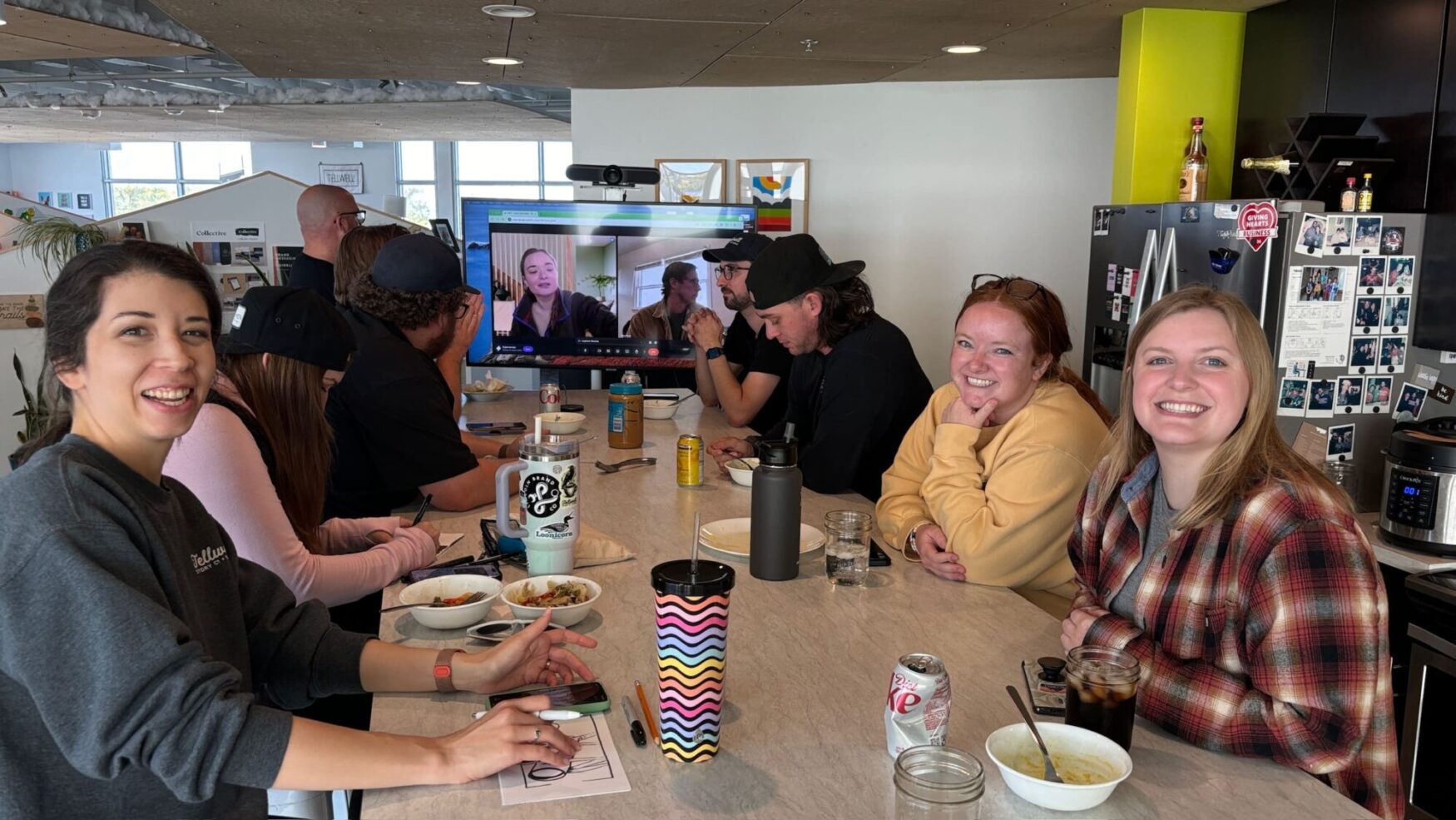 A group of people sitting around a table in a casual office setting, enjoying food and drinks while watching a virtual meeting on a large screen. Several individuals are smiling and engaged in conversation, creating a warm, collaborative atmosphere. The background includes office decor, personal photos, and a screen on which two people are engaged with the group virtually, adding a relaxed and personal touch to the workspace.