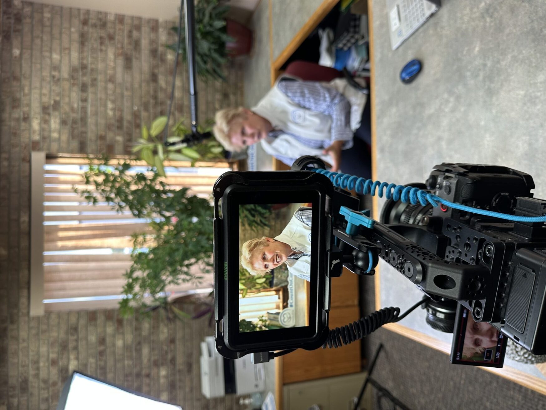 Camera focused on a woman speaking during an interview, shown through the monitor of a professional filming setup. The woman wears a light-colored vest and sits in an office environment with plants in the background.