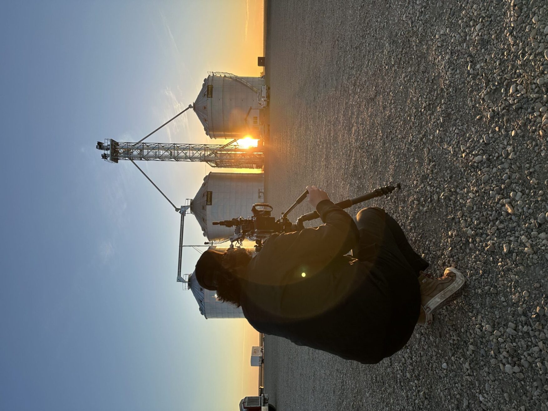 Person crouching with a camera on a tripod, capturing a scene of large grain silos at sunrise or sunset. The sun is partially visible between the silos, casting a warm glow across the landscape.