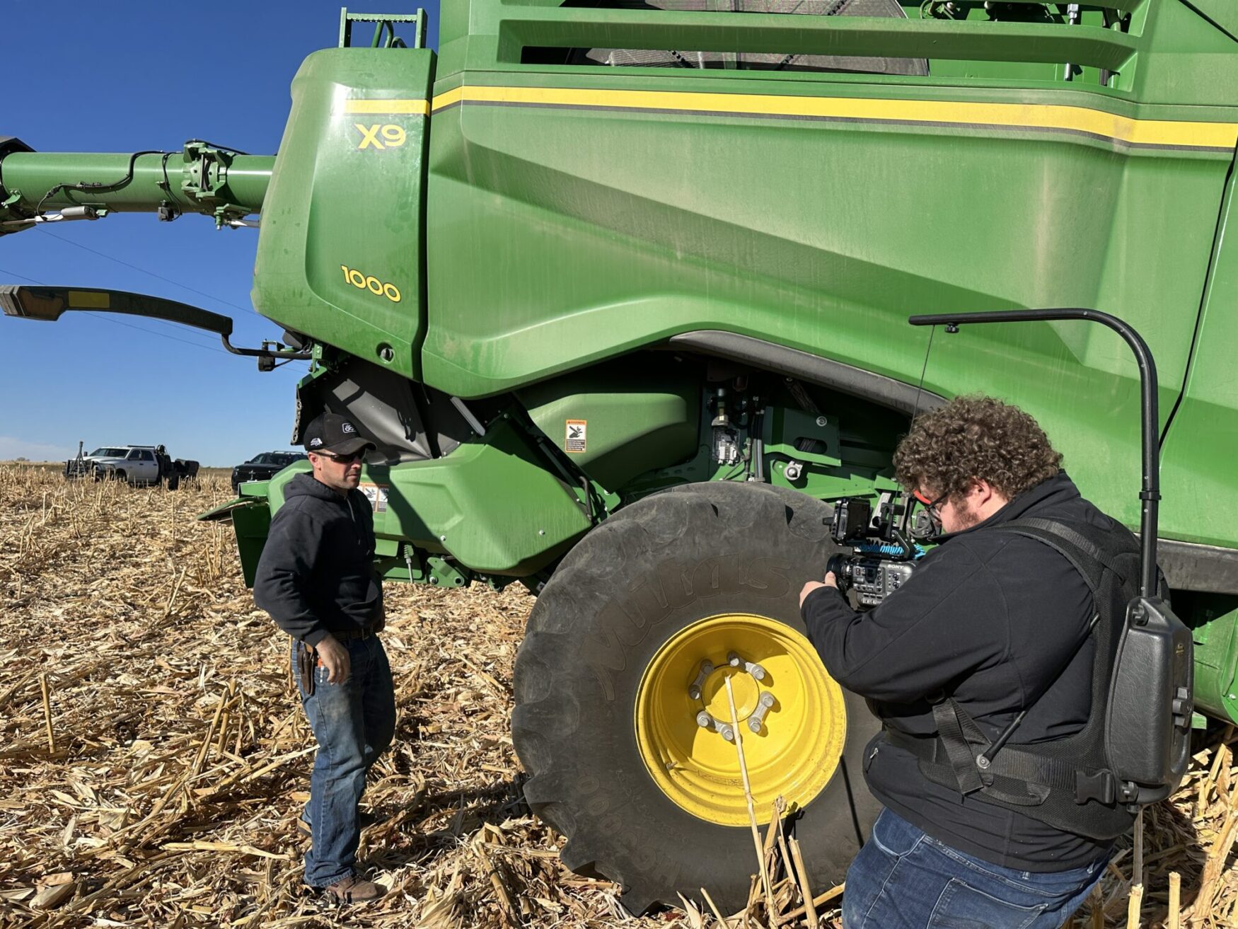 A film crew member wearing headphones is filming a man standing next to a large green John Deere tractor in a cornfield. The sun shines brightly on the field, with the focus on the large tractor wheel and the camera setup.